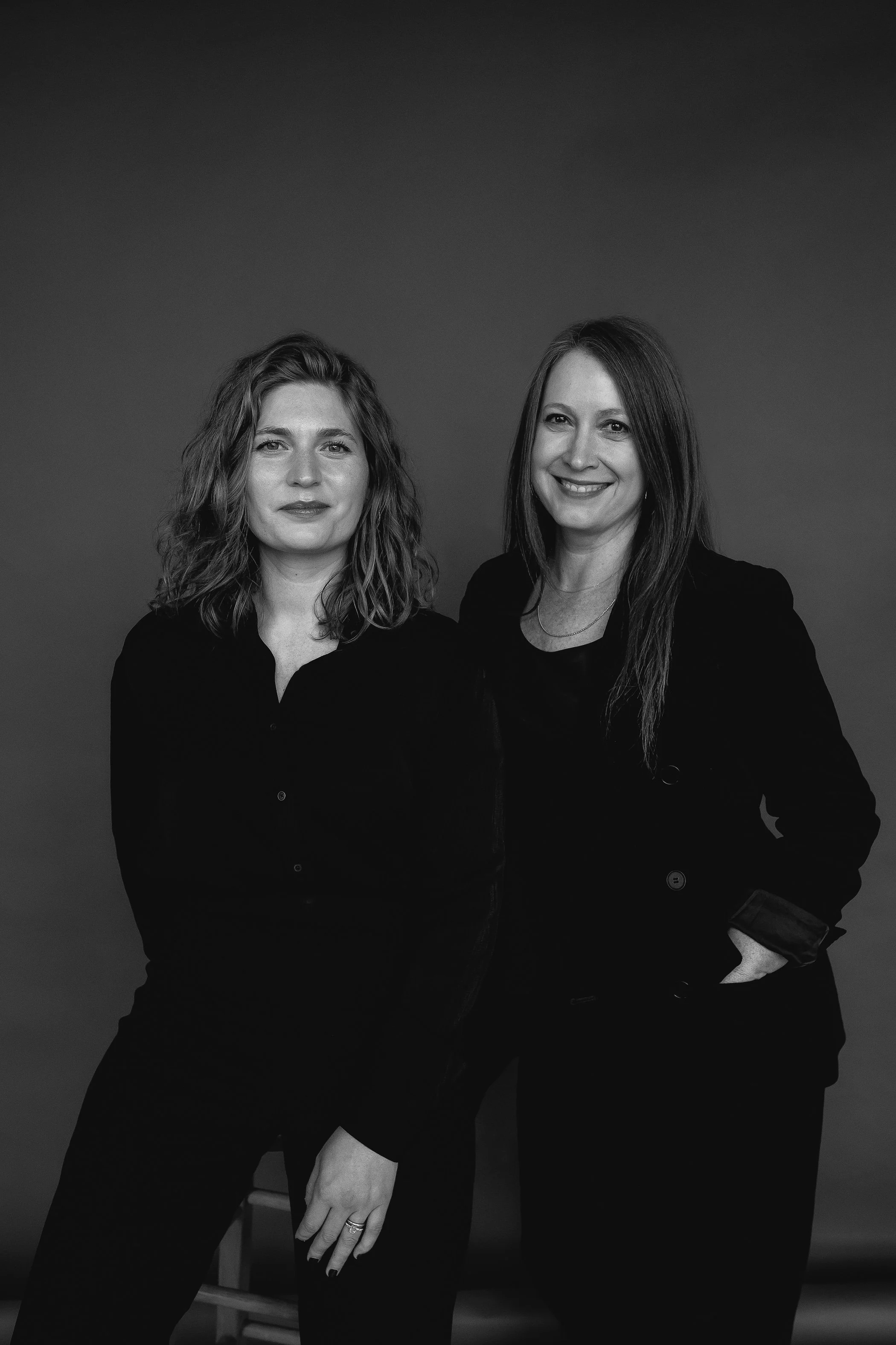 Two women standing side by side against a plain background, dressed in dark clothing, one with curly hair and the other with straight hair, smiling at the camera.