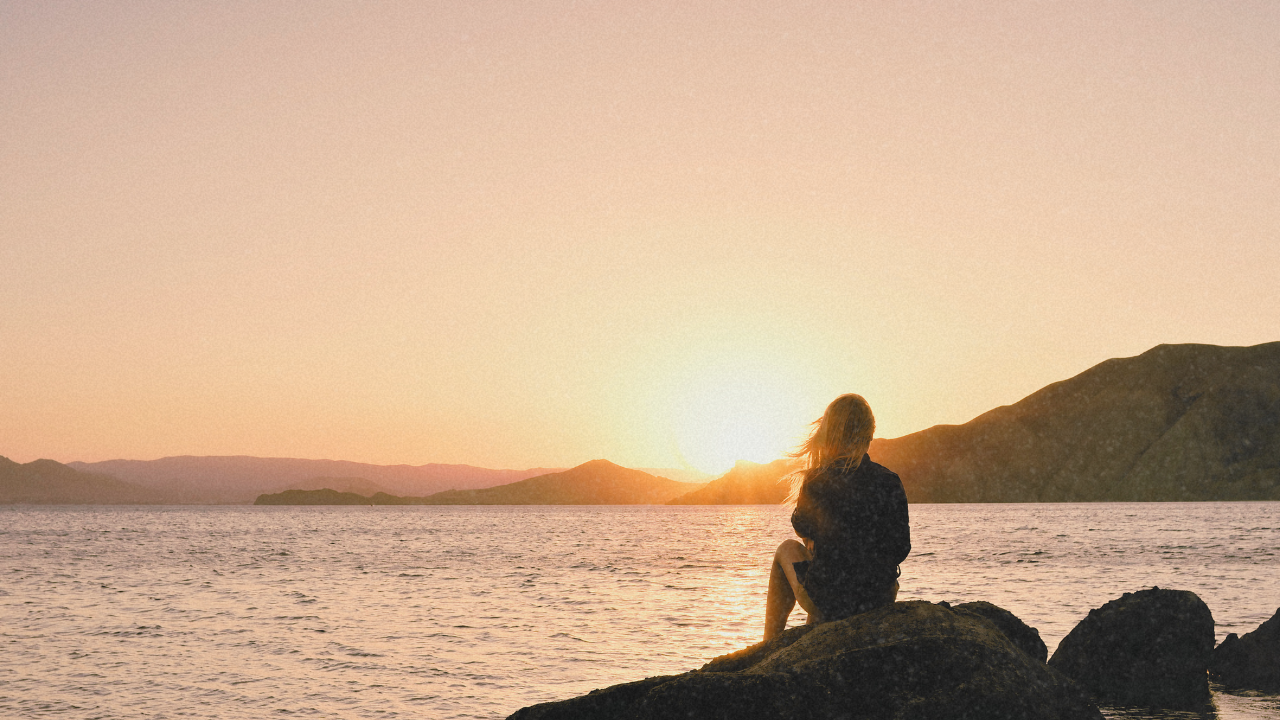 Woman sitting on rocks watching the sunrise over the ocean, representing healing and finding herself again.