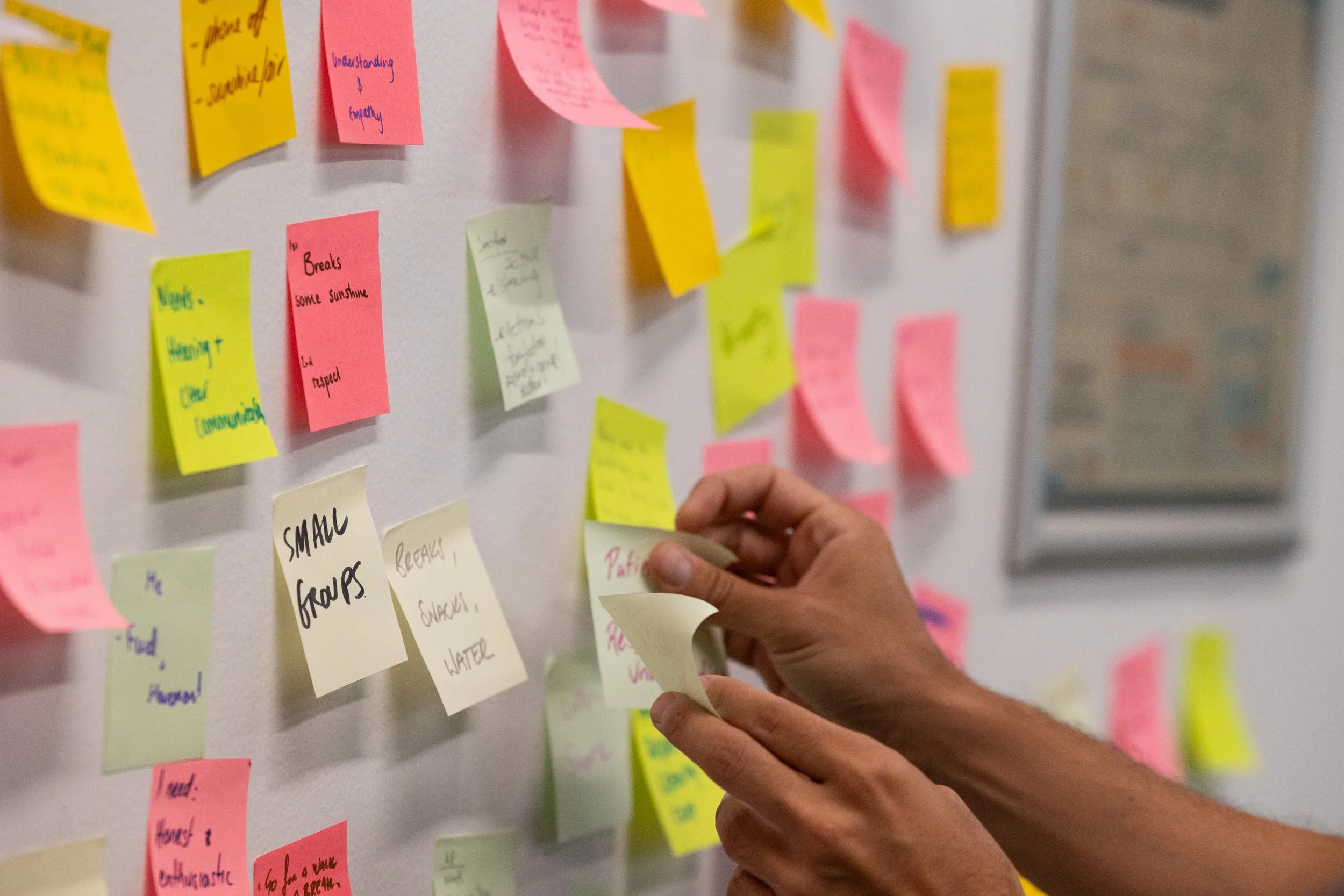 A pair of hands place a sticky note onto a wall covered in brightly coloured sticky notes.