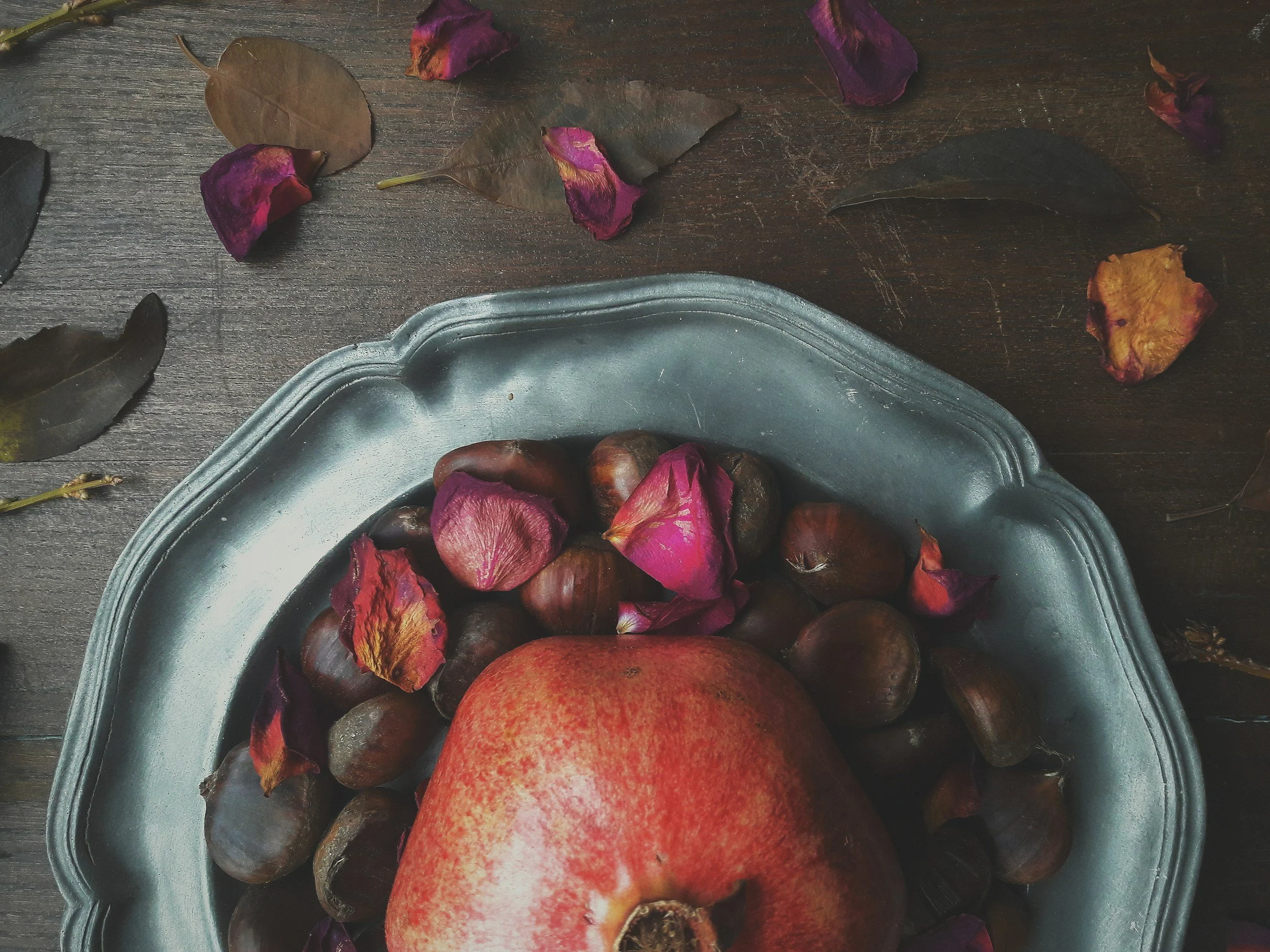 A silver tray filled with pomegranate seeds and dried rose petals on a wooden table, with additional rose petals and leaves scattered around.