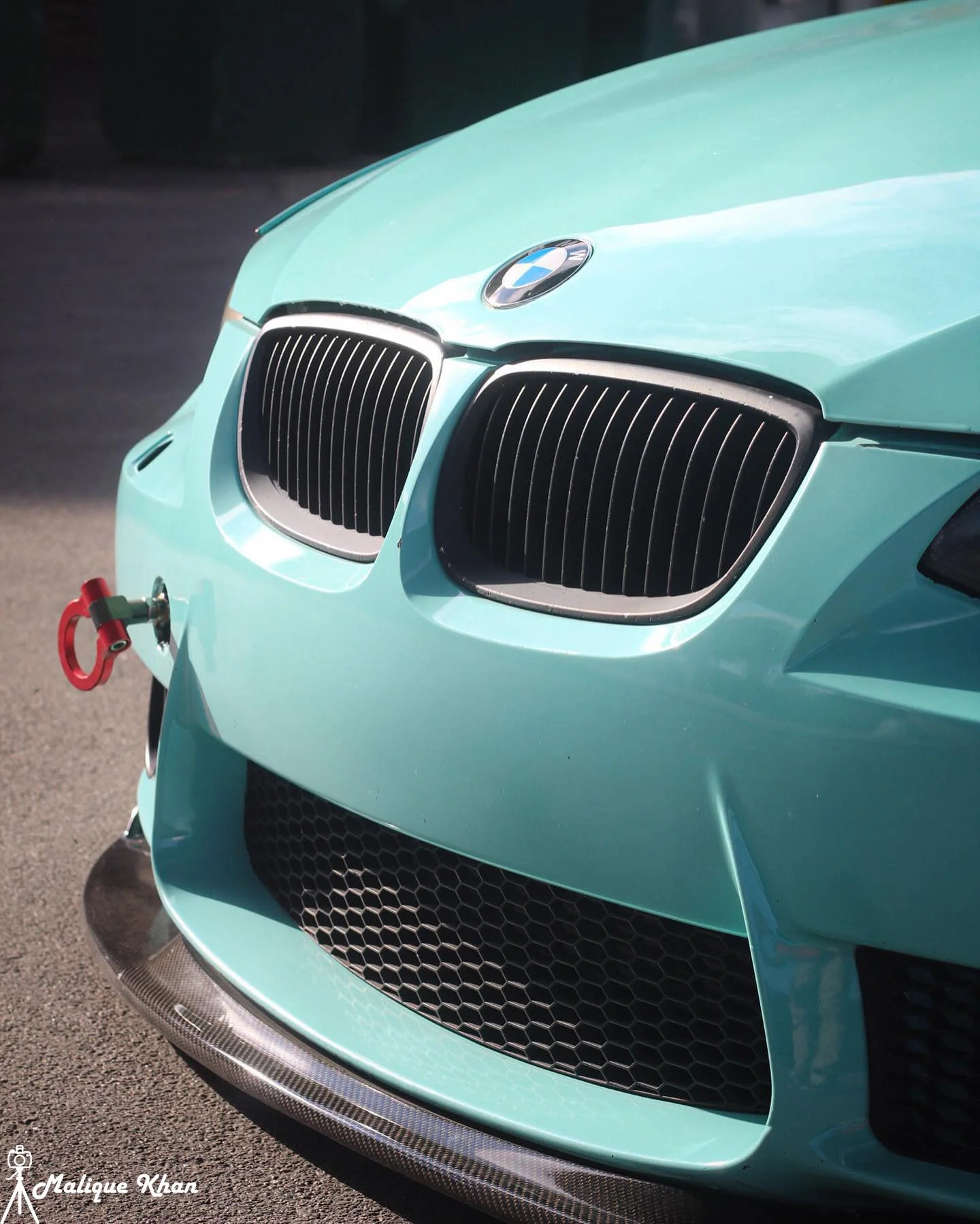 Close-up of a turquoise BMW car's front grill, hood, and bumper, with a red tow hook on the left side.