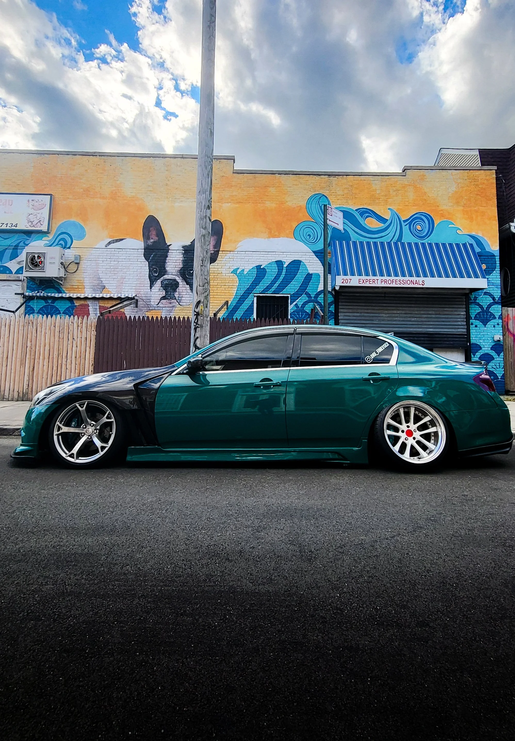 A low-profile, dark green custom car parked on the street in front of a colorful mural featuring a French Bulldog puppy and ocean waves, with a building and cloudy sky in the background.