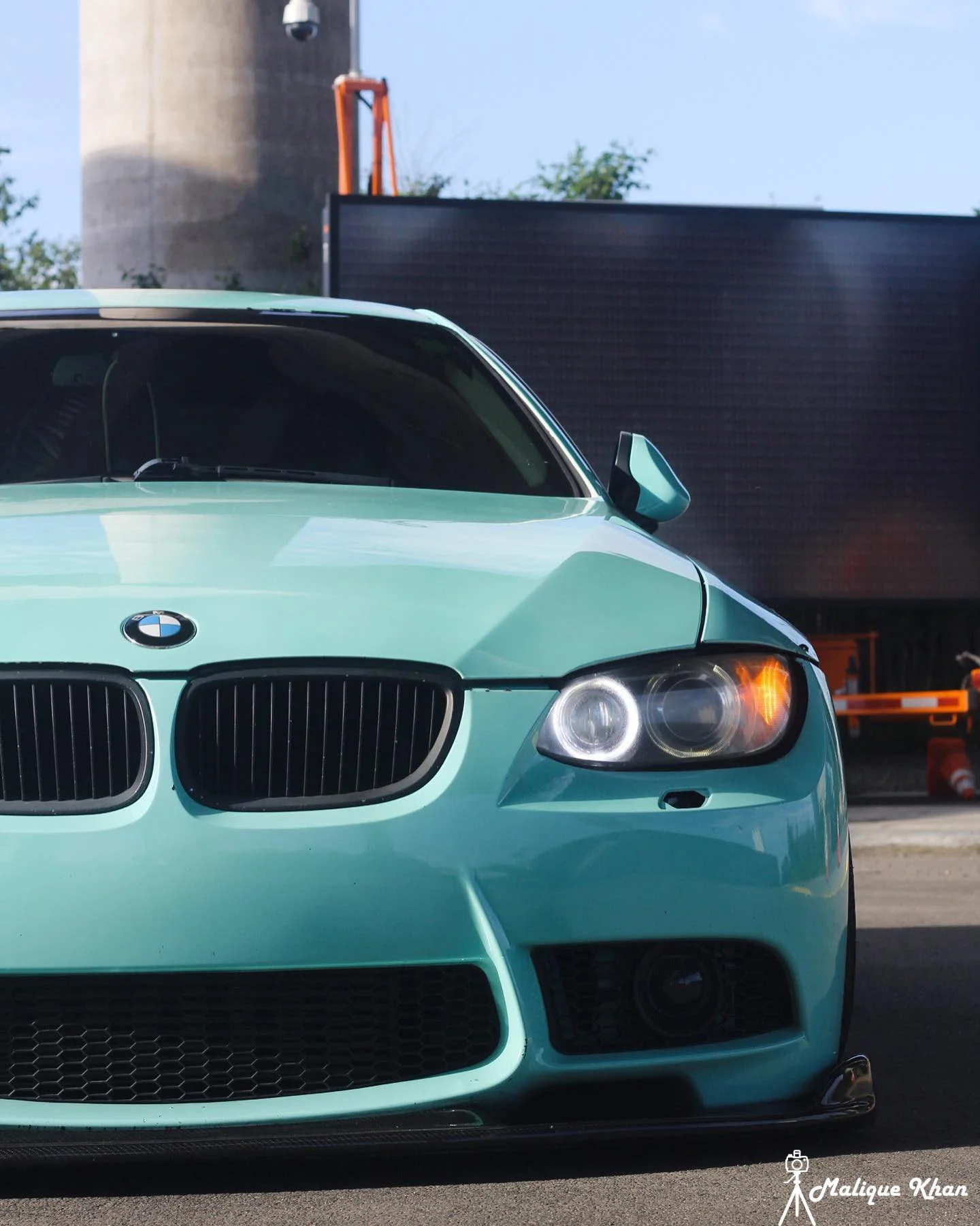 Front view of a mint green BMW car parked outdoors with a black screen in the background and industrial structures visible in the distance.