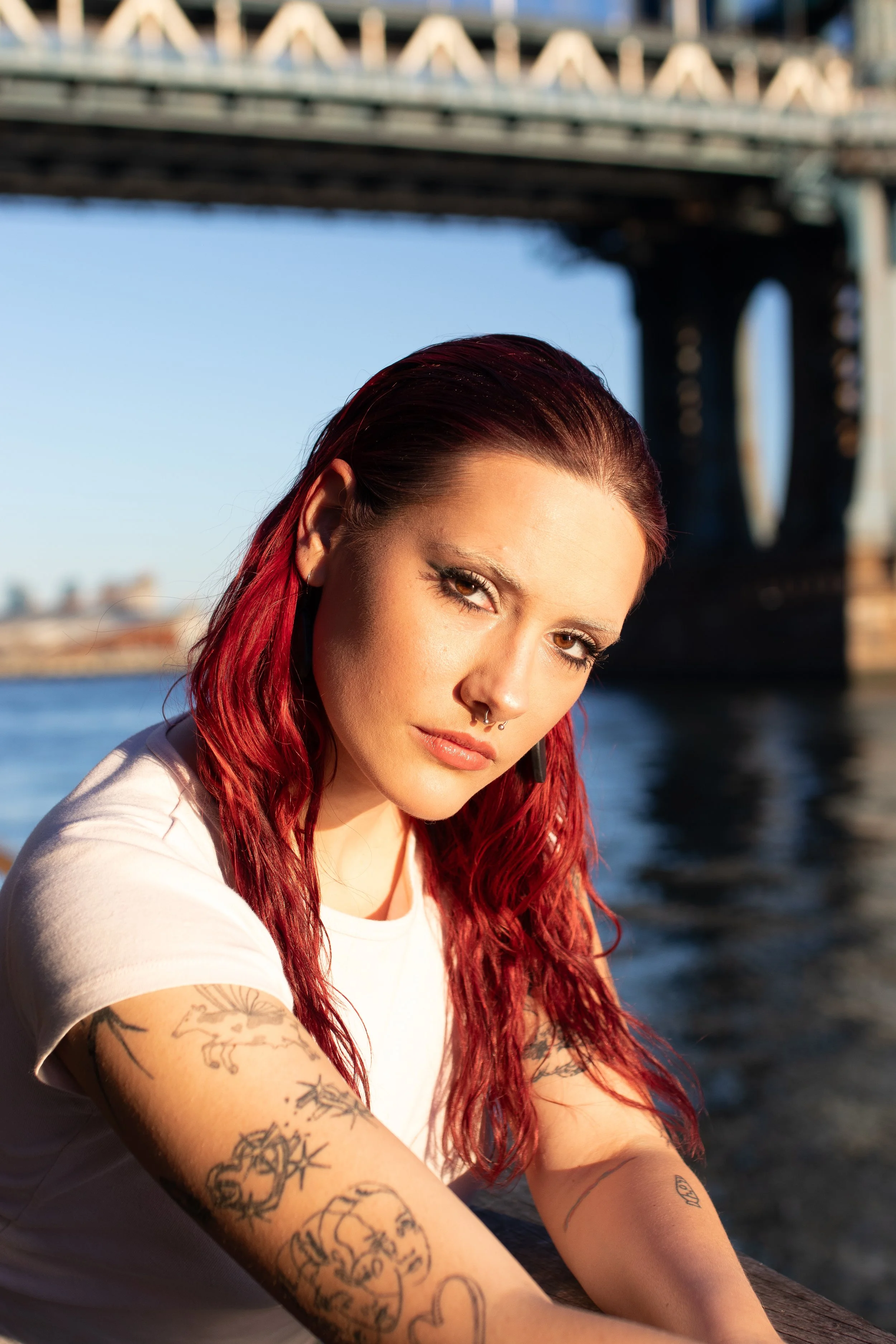 Portrait of a woman with red hair, tattoos on her arms, and piercings, sitting outdoors near a river with a bridge in the background during sunset.