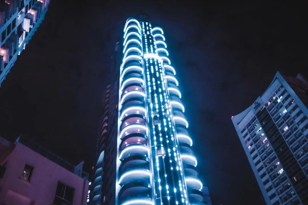 Night view of a tall skyscraper with illuminated blue and white LED lights, surrounded by other high-rise buildings.