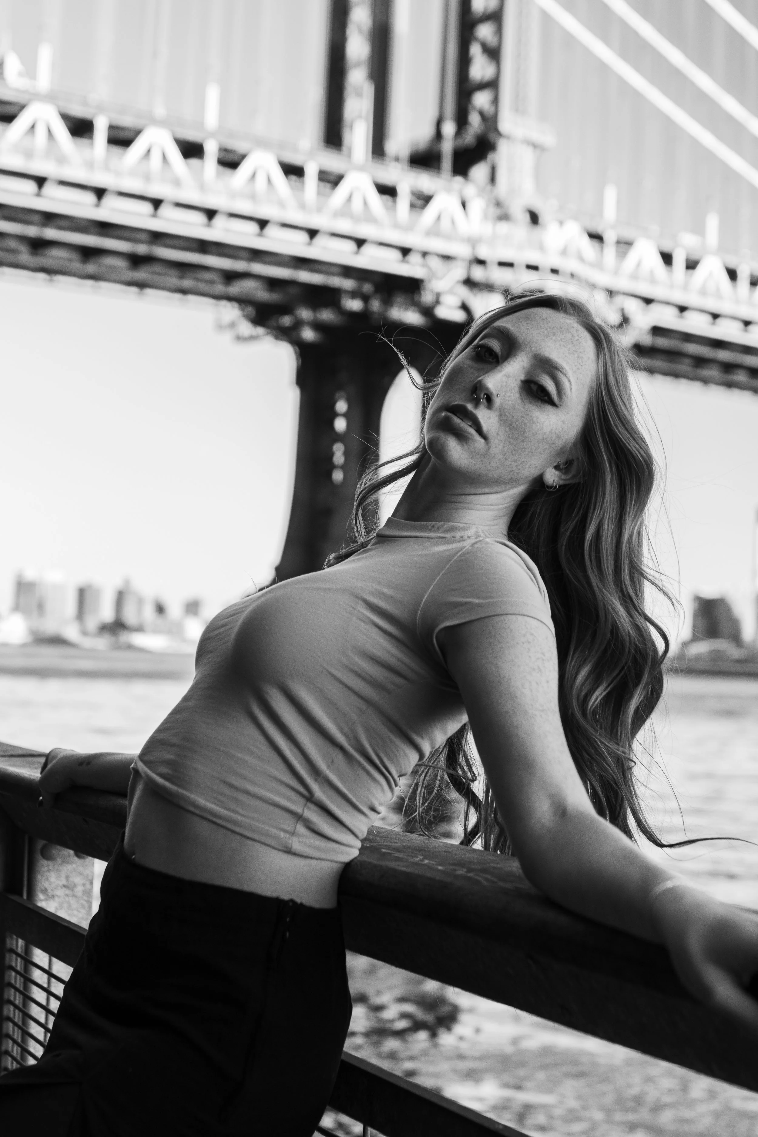 A woman with long hair and a nose piercing leaning against a wooden railing by a river, with a bridge in the background, in black and white.