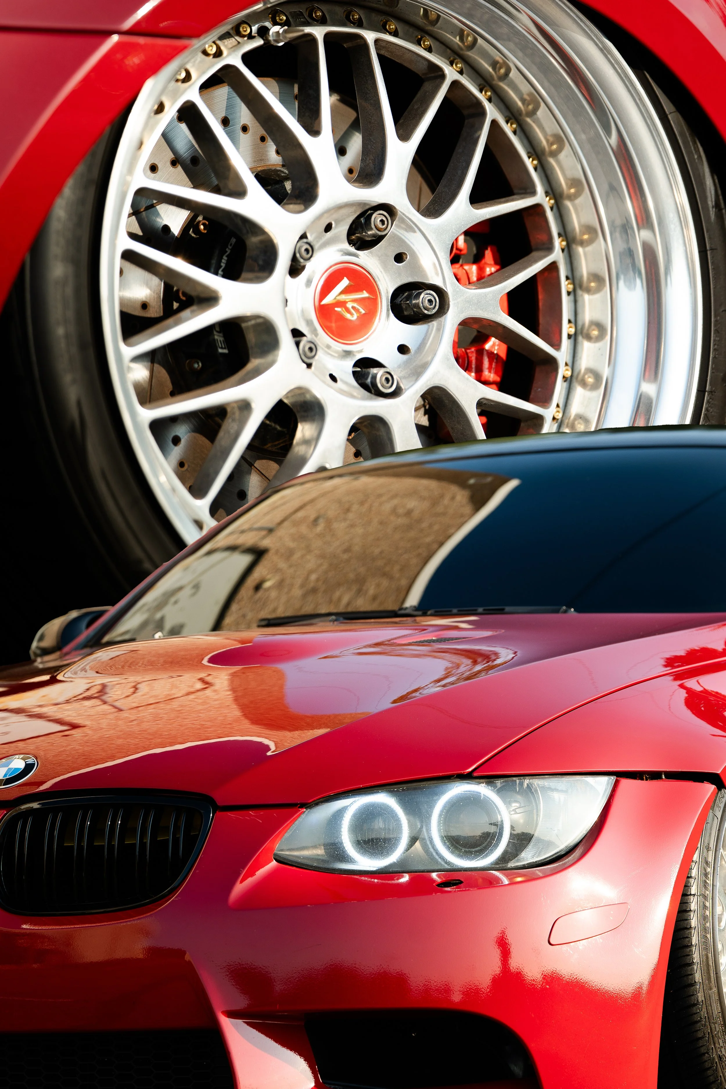 Close-up of a red BMW sports car with glowing headlights, showing part of the front and a reflection of the sky on the hood, with a blurred background.