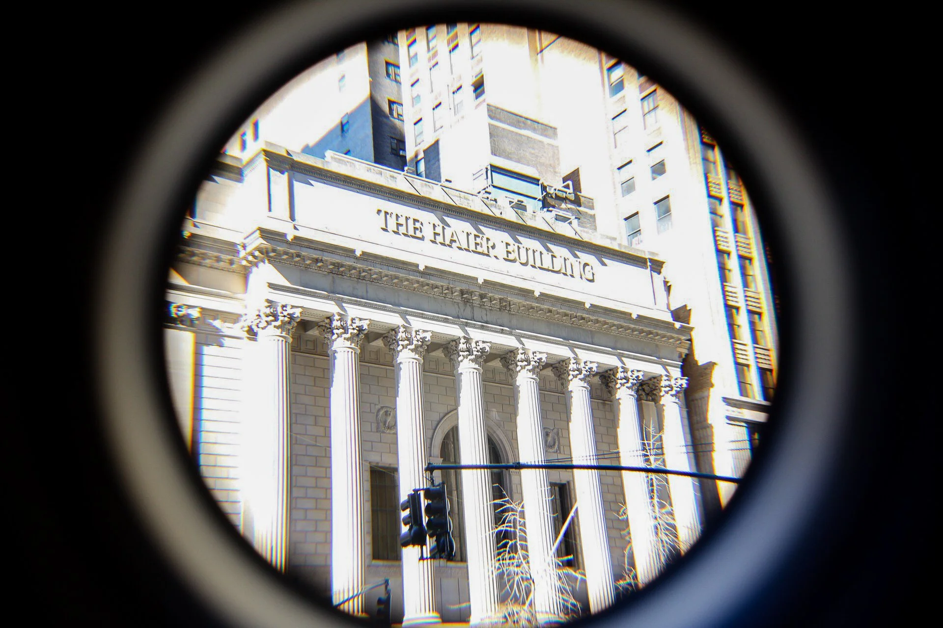 A view of a historic building called 'The Hayden Building' seen through a circular peephole, with tall modern buildings in the background and a traffic light in the foreground.