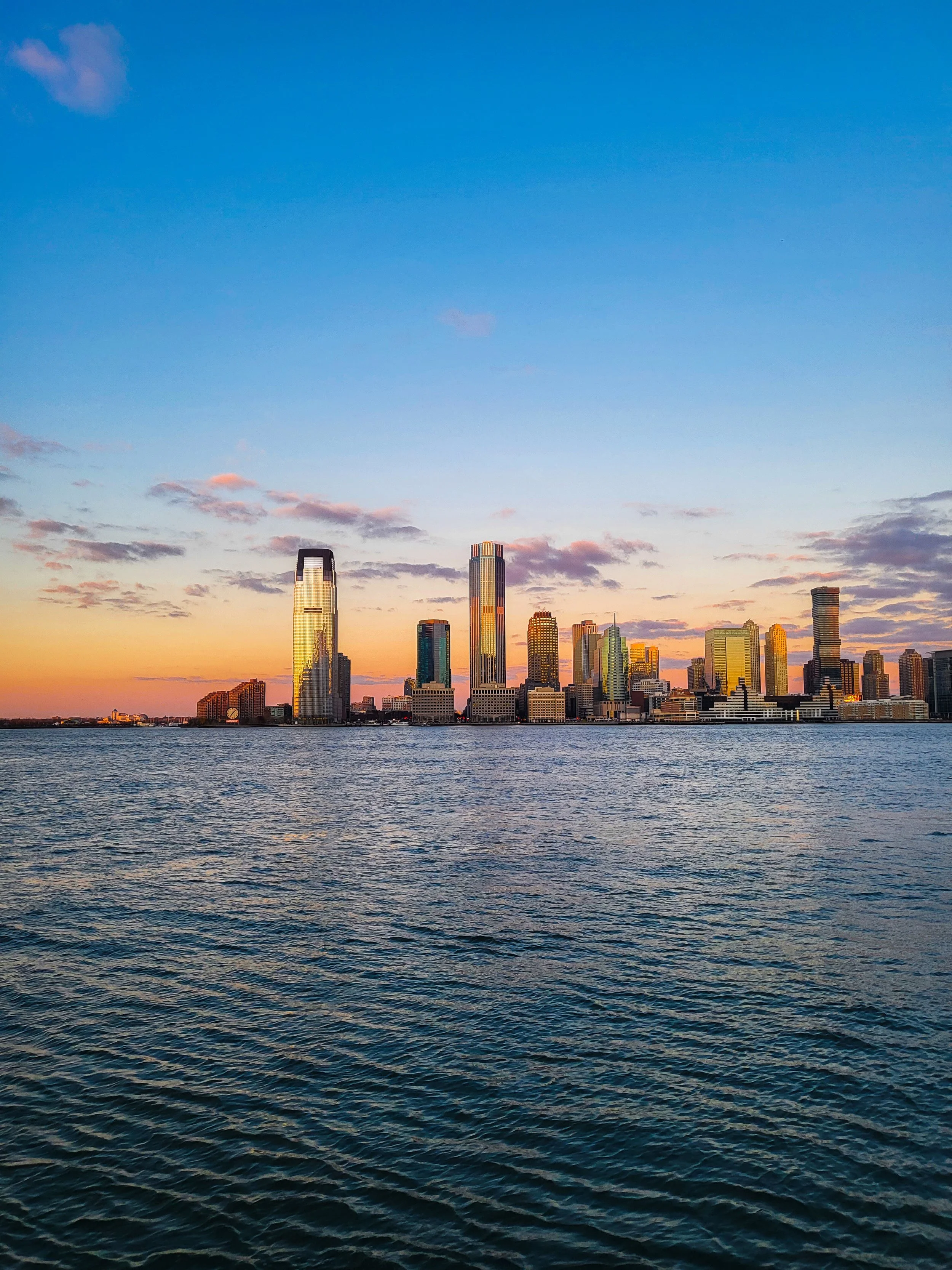 Sunset view of a city skyline with tall skyscrapers along the waterfront, with colorful sky and water reflections.