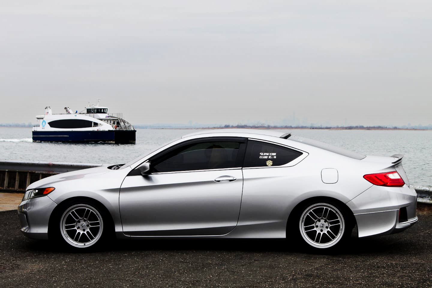 A silver coupe car parked on a paved area near a body of water with a ferry in the background under an overcast sky.