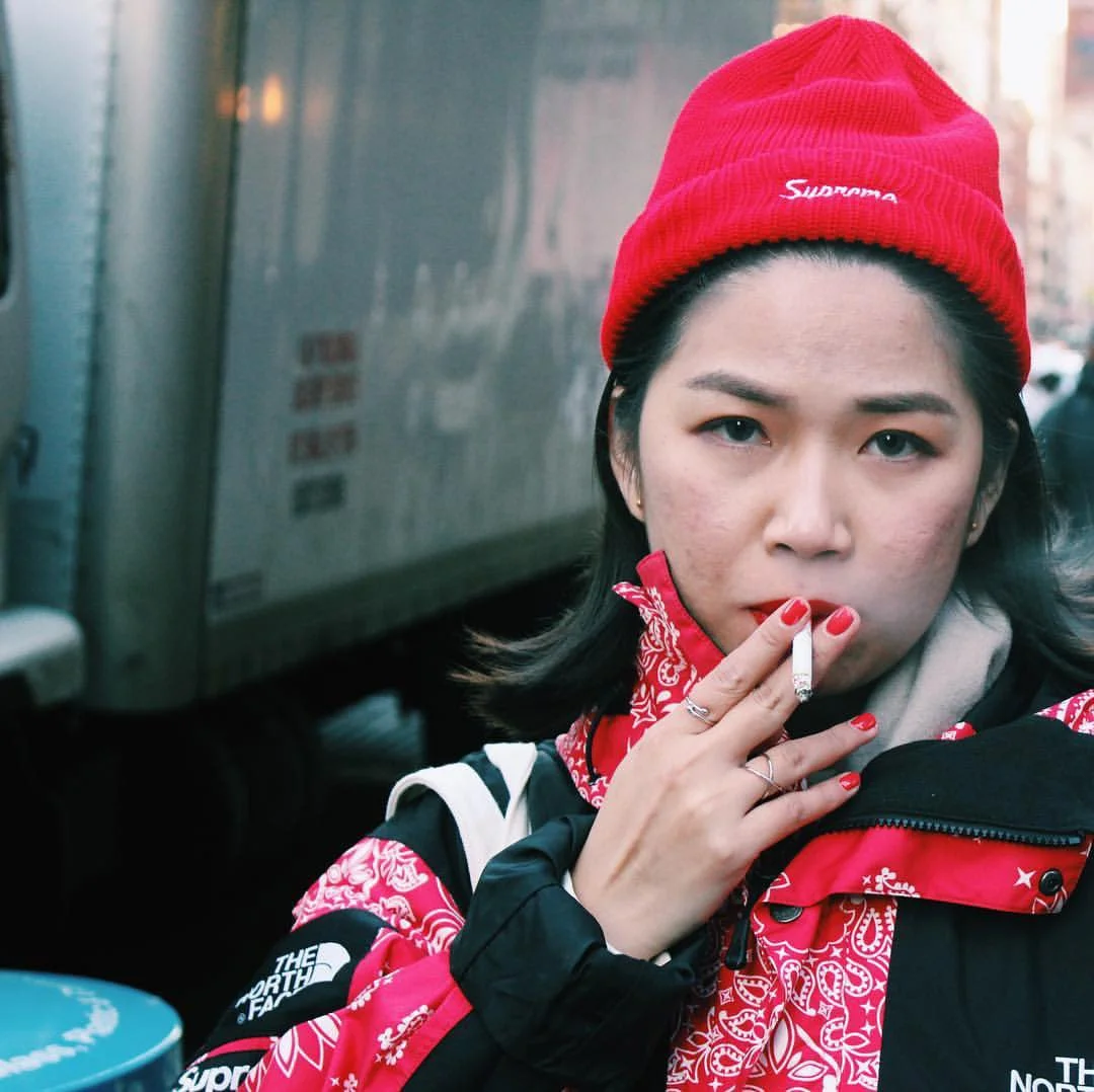 Young woman with dark hair and light skin wearing a red beanie, red bandana around her neck, and a black and red jacket with logos, holding a cigarette near her lips, outdoors in an urban setting.