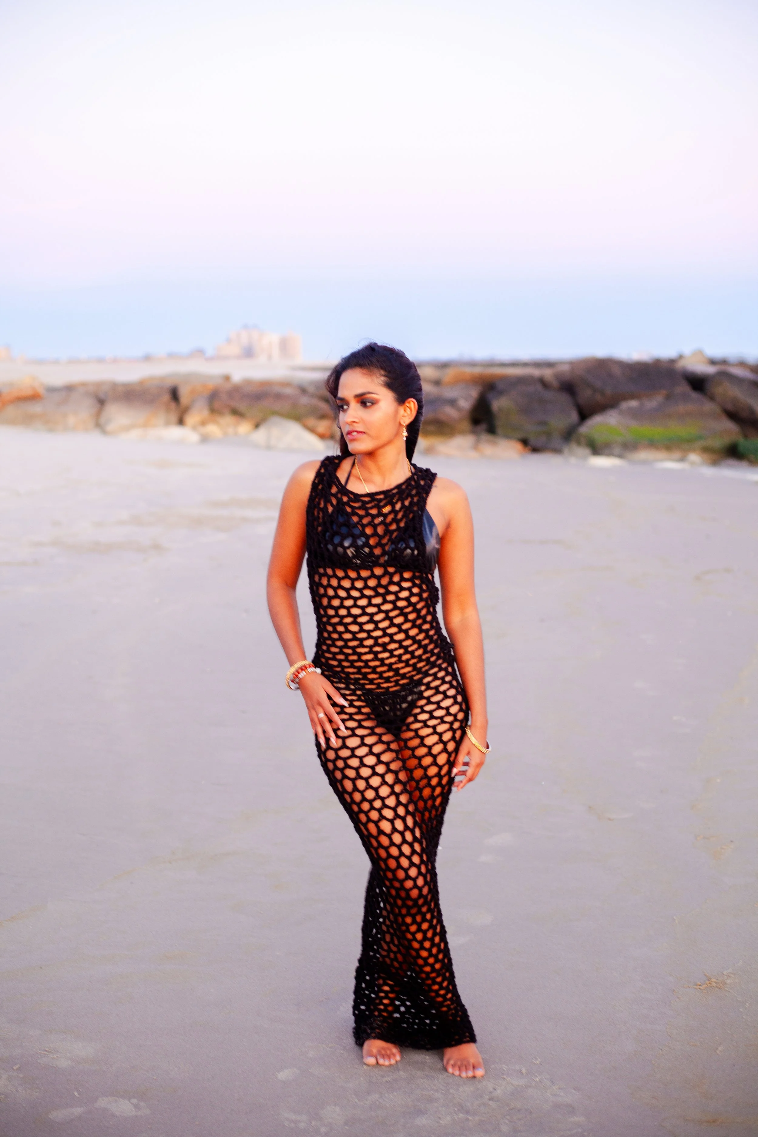 A woman in a black crochet dress standing on a beach with rocks in the background during sunset.