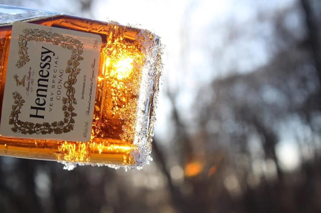 A bottle of Hennessy Very Special Cognac ice-covered in frost, with the sun shining through it, outdoors in a winter setting with blurred trees in the background.
