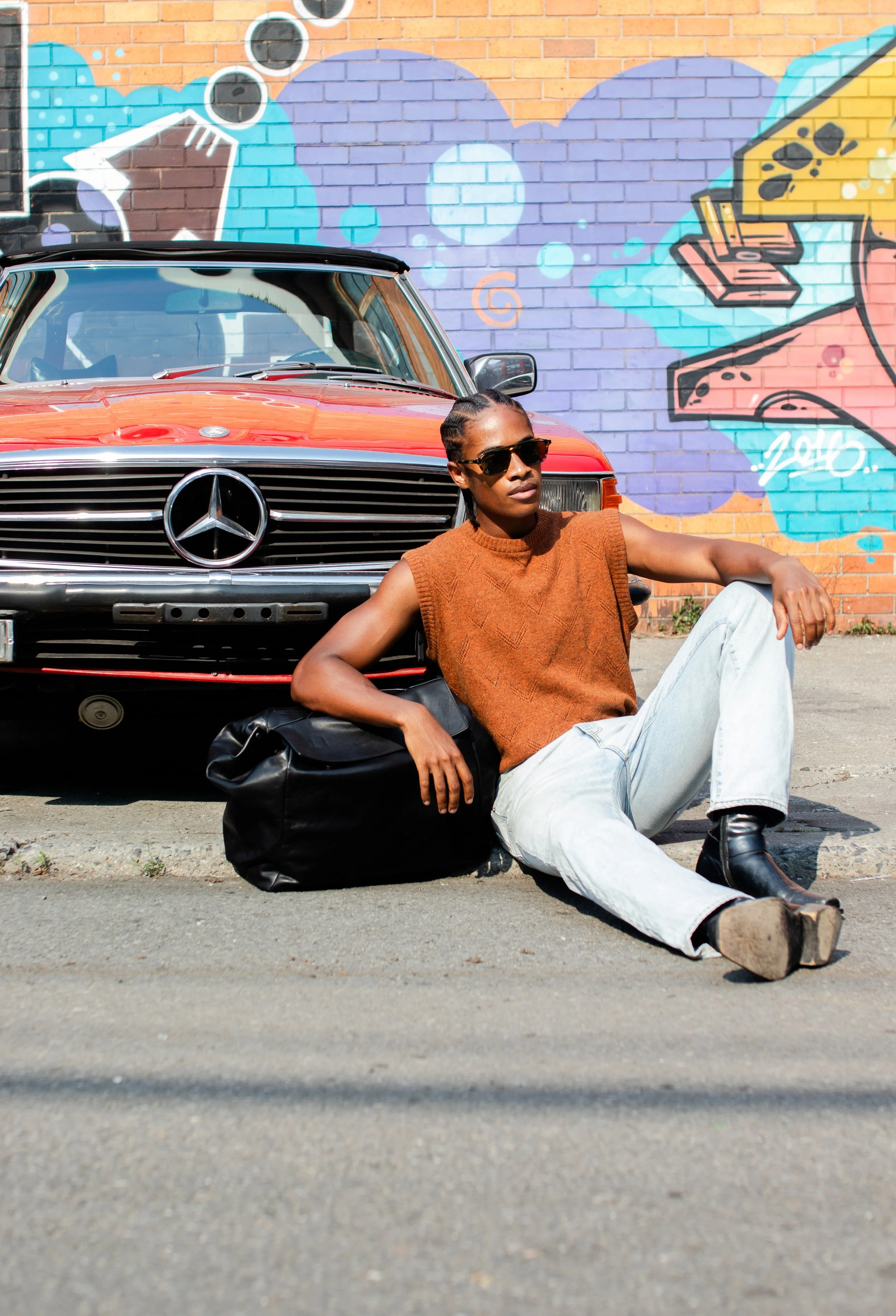 A woman with braided hair, wearing sunglasses, a sleeveless orange sweater, and light-colored jeans, leaning against a black bag and a red vintage Mercedes-Benz car, in front of a colorful graffiti wall.