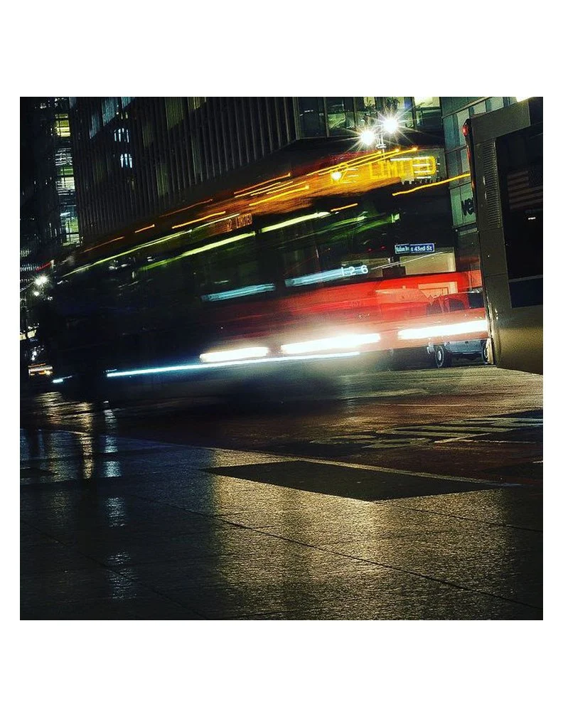 A city street at night with a bus and its light trails from motion blur, reflecting on wet pavement.