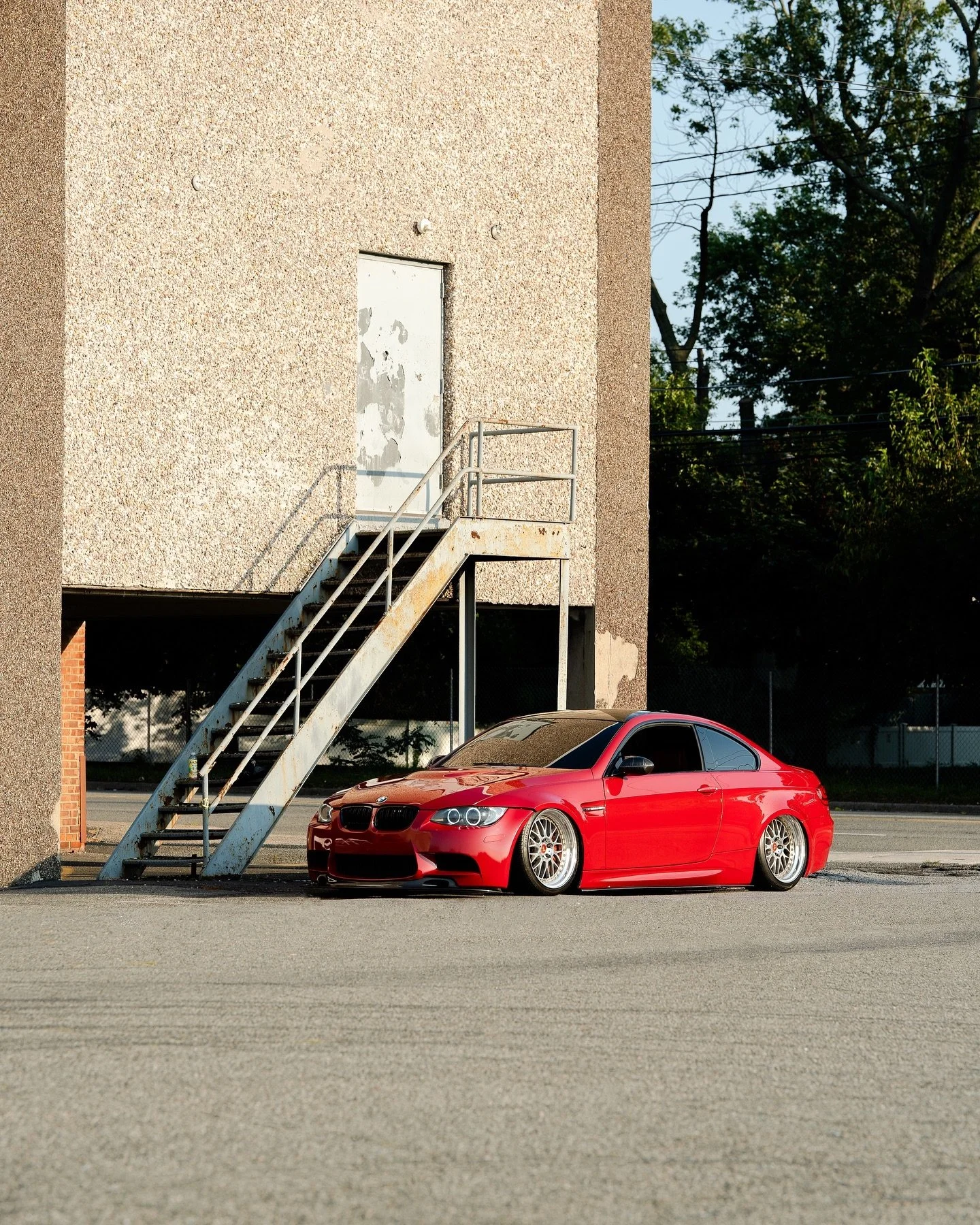 A red sports car parked next to a building with a metal staircase and a closed door. The car is low to the ground with custom wheels, reflecting sunlight, and is positioned on a paved surface with trees and a fence in the background.