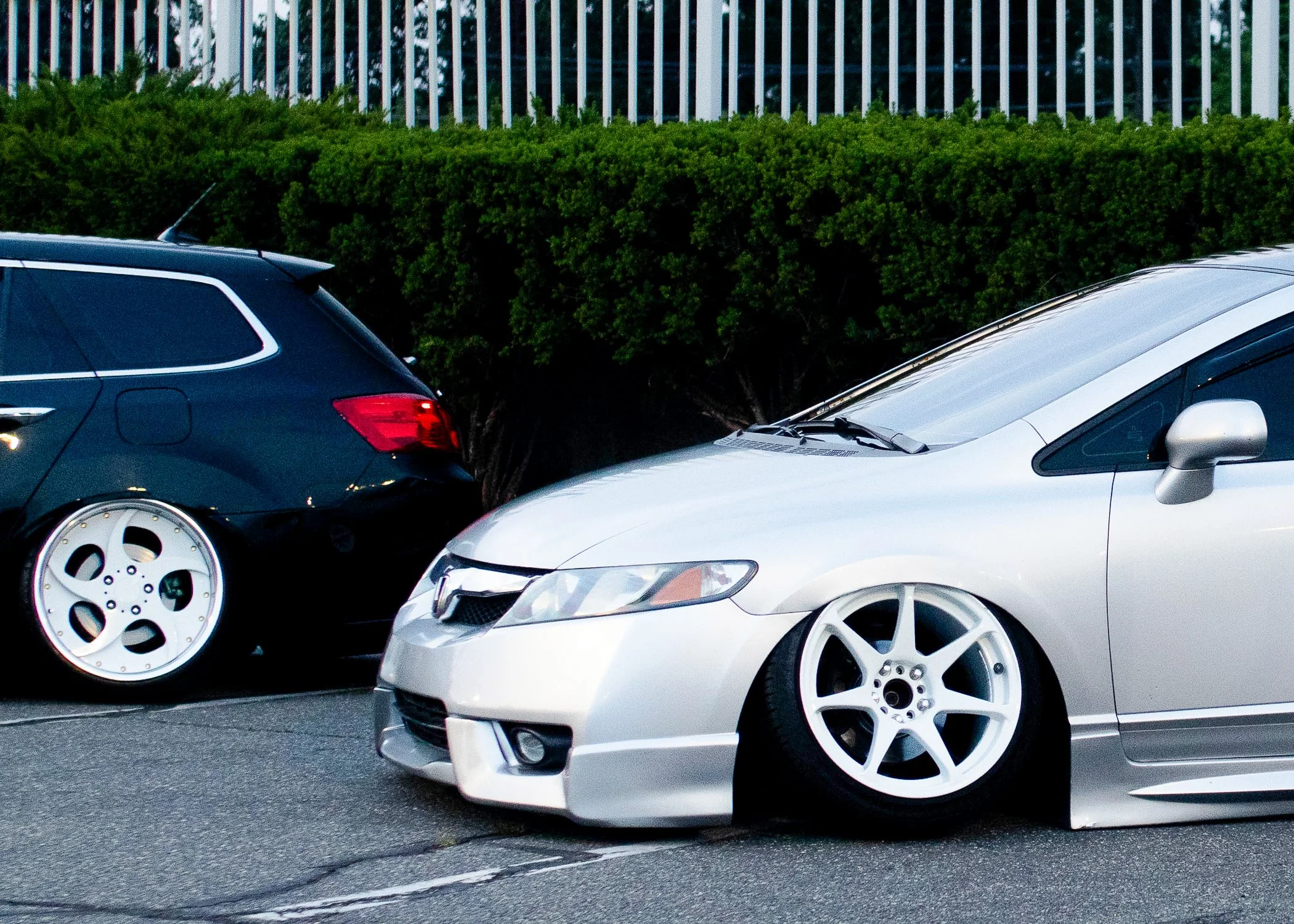 A silver car with custom white wheels parked next to a black SUV with custom white wheels in a parking lot, with a metal fence and bushes in the background.