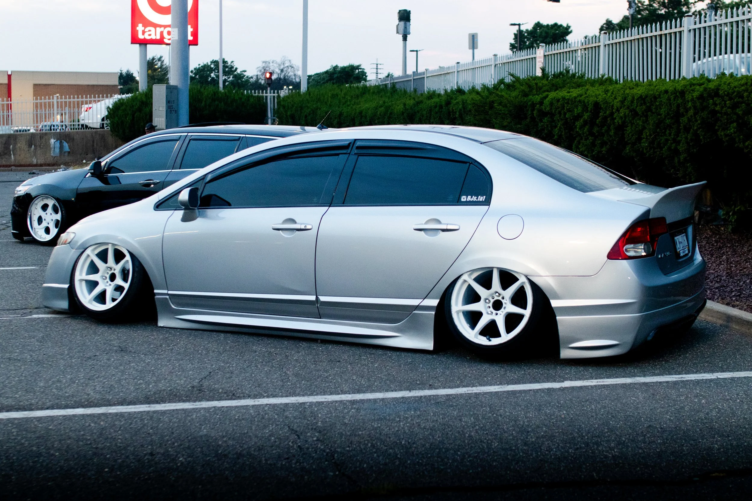 A silver modified car with lowered suspension and custom white wheels parked in a parking lot next to a black car. There is a Target store and a fence with bushes in the background.