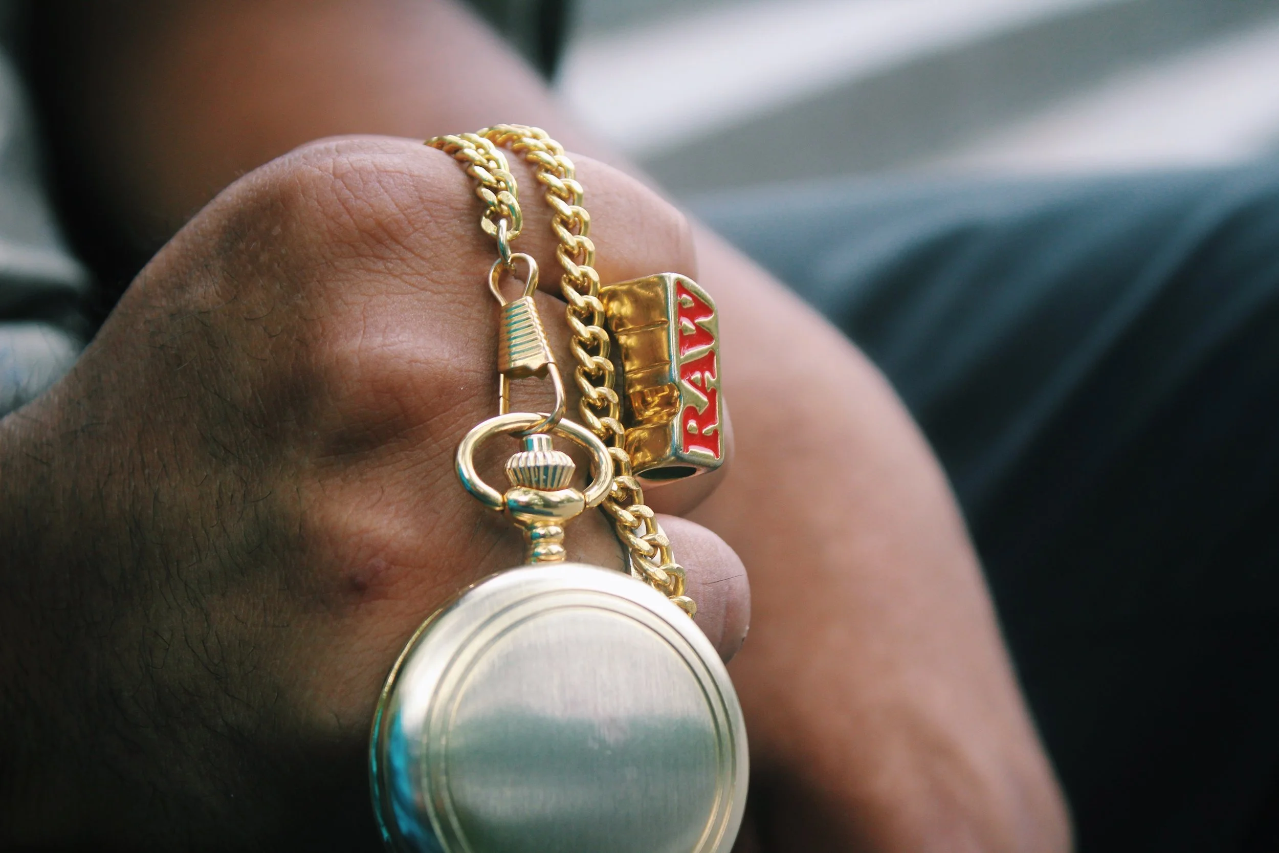 Close-up of a hand with a gold chain, a ring with the word 'RAY' in red, and a round metal object attached to the chain.