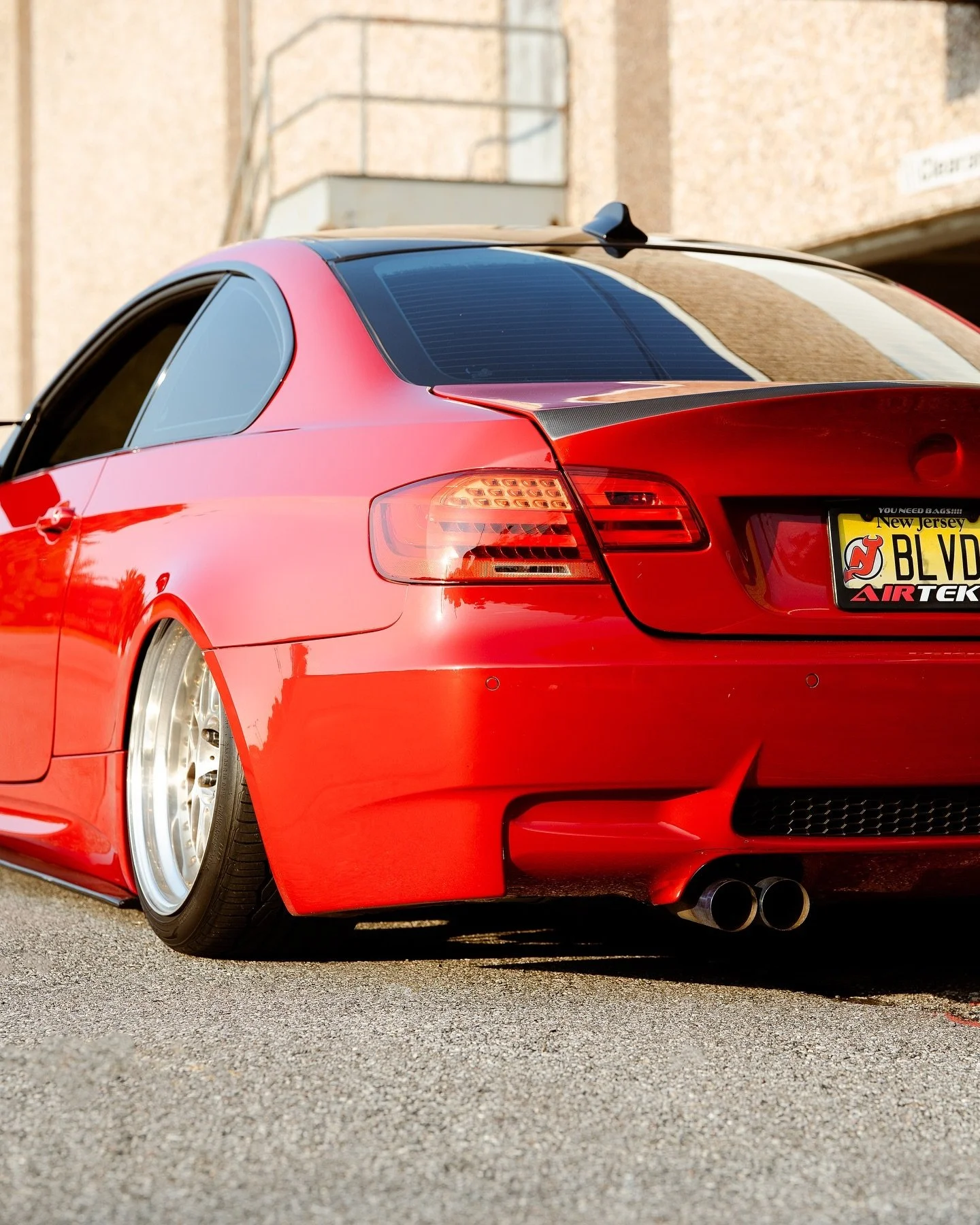 Red sports car with aftermarket wheels and dual exhaust pipes parked on a street, showing the rear and driver side, with a yellow New Jersey license plate and a black carbon fiber spoiler on the trunk.