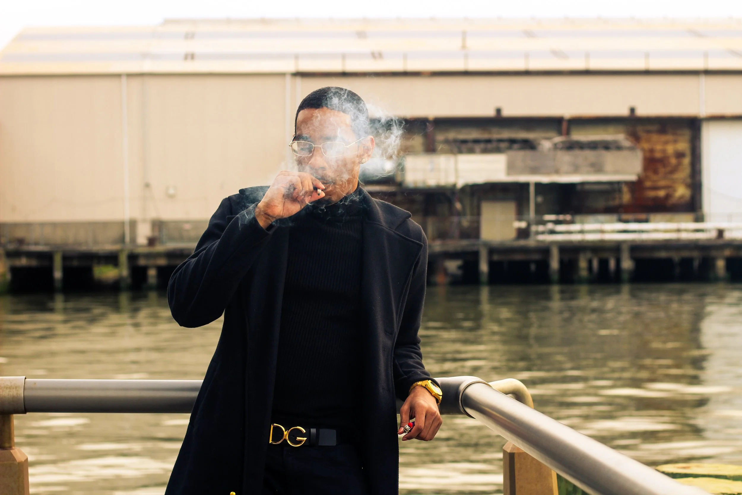 A man with glasses and a gold watch is smoking a cigarette by the water near a metal railing, with industrial buildings in the background.