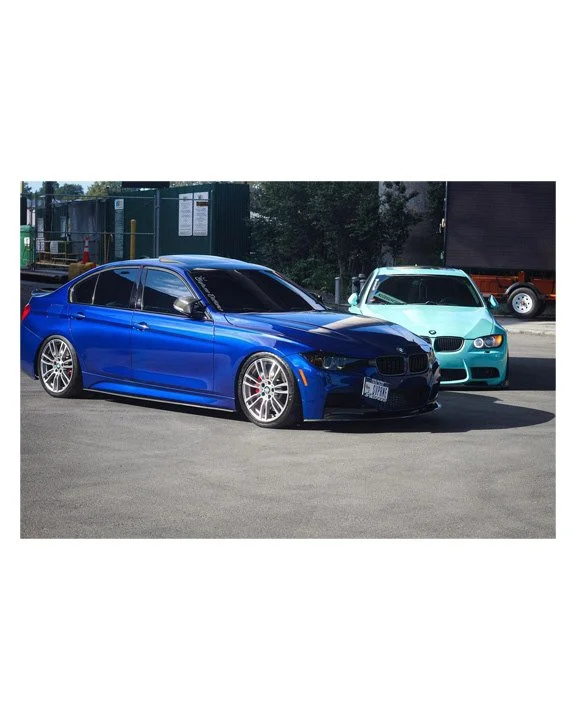 Two parked cars, a blue BMW sedan in the foreground and a light blue BMW coupe in the background, on a city street with industrial fencing and trees.