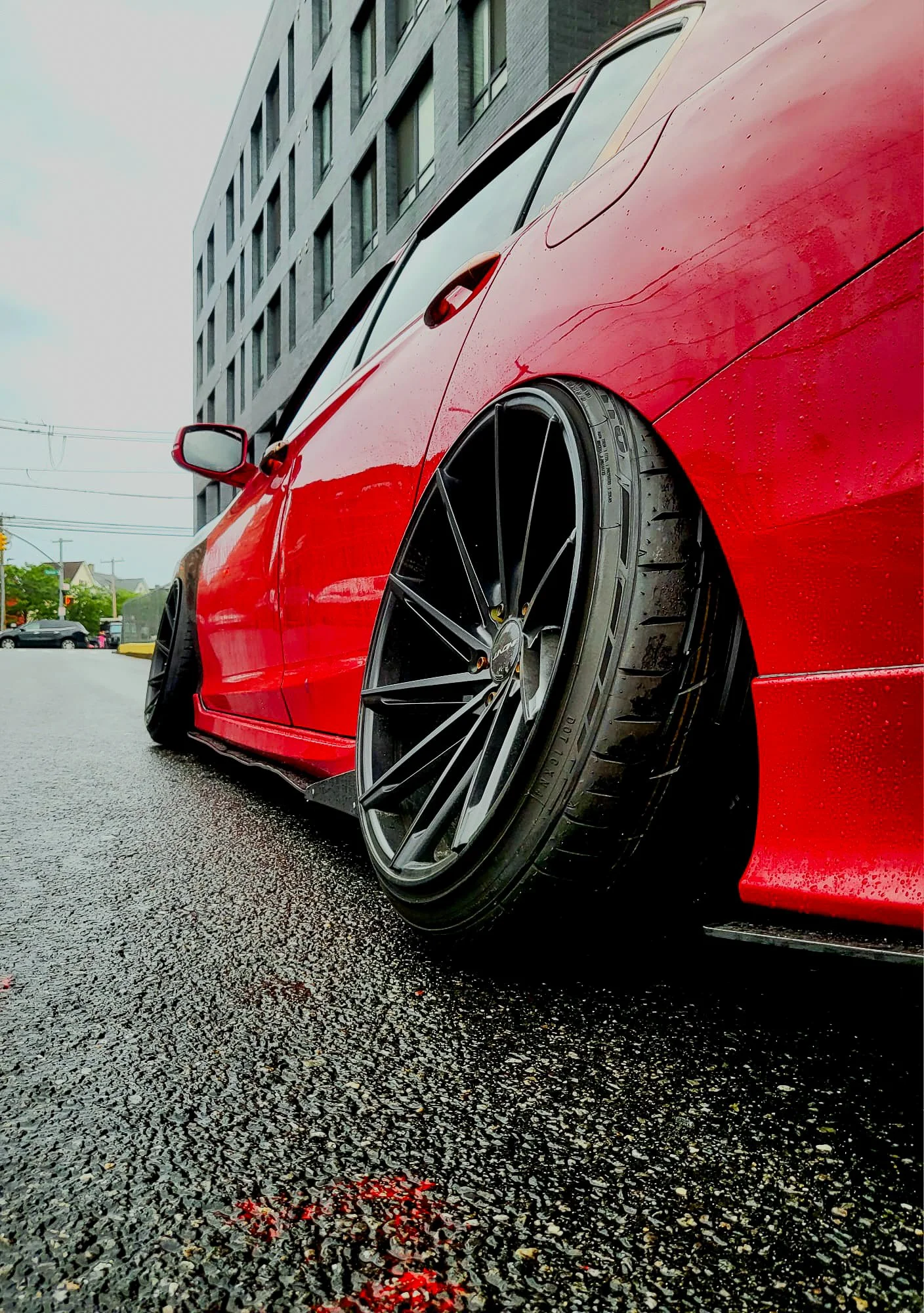 Close-up of a red sports car with black rims and low-profile tires parked on wet asphalt, with a modern multi-story building in the background.