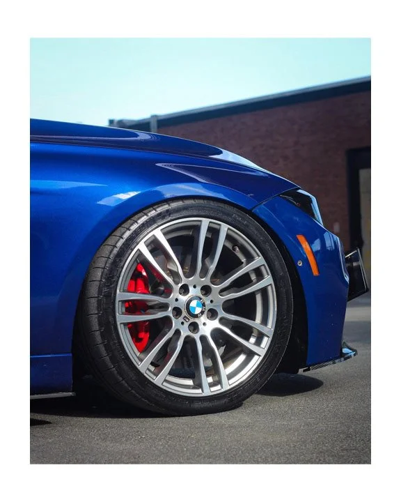 Close-up of a blue BMW car's front wheel with a silver alloy rim and red brake caliper, parked on the pavement near a brick building.