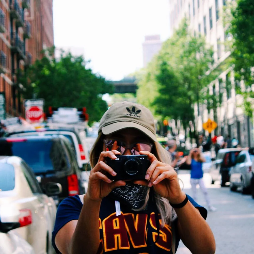 Person taking a photo on a city street with trees and parked cars.