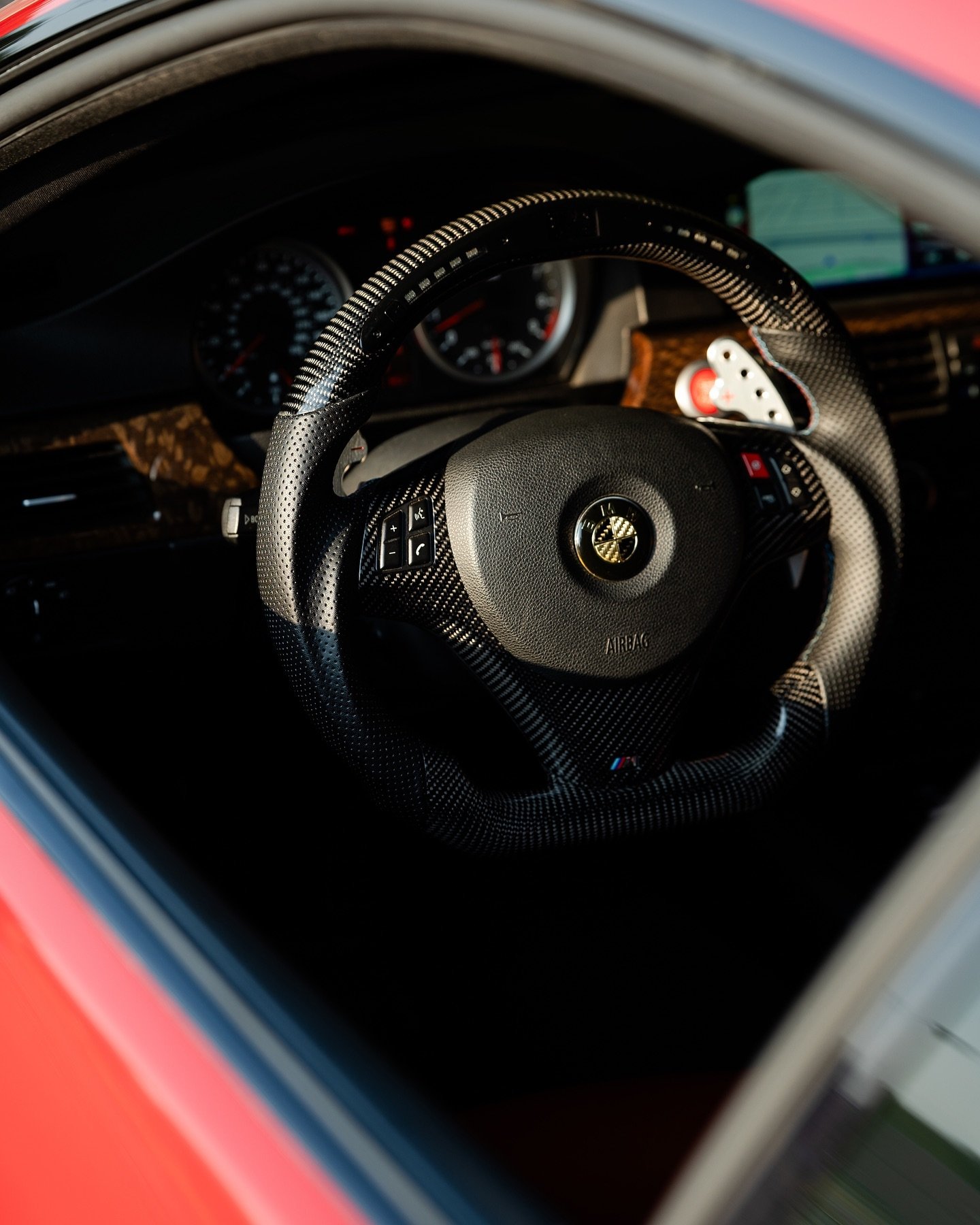Close-up of a car's interior steering wheel with a carbon fiber covering, mounted on a dashboard with gauges and a navigation screen in background.