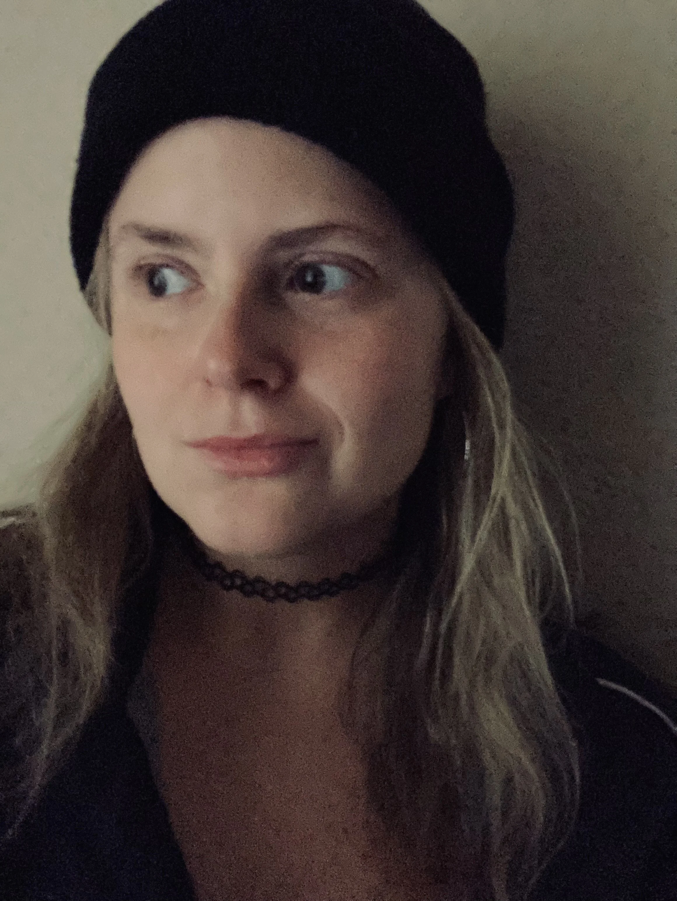 A woman with wavy blonde hair, wearing a black beanie, dark clothing, and a black choker necklace, posing against a neutral background.