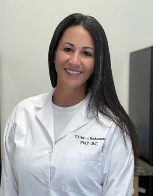 A woman with long dark hair smiling, wearing a white coat with a name tag, standing in an office or clinic setting.
