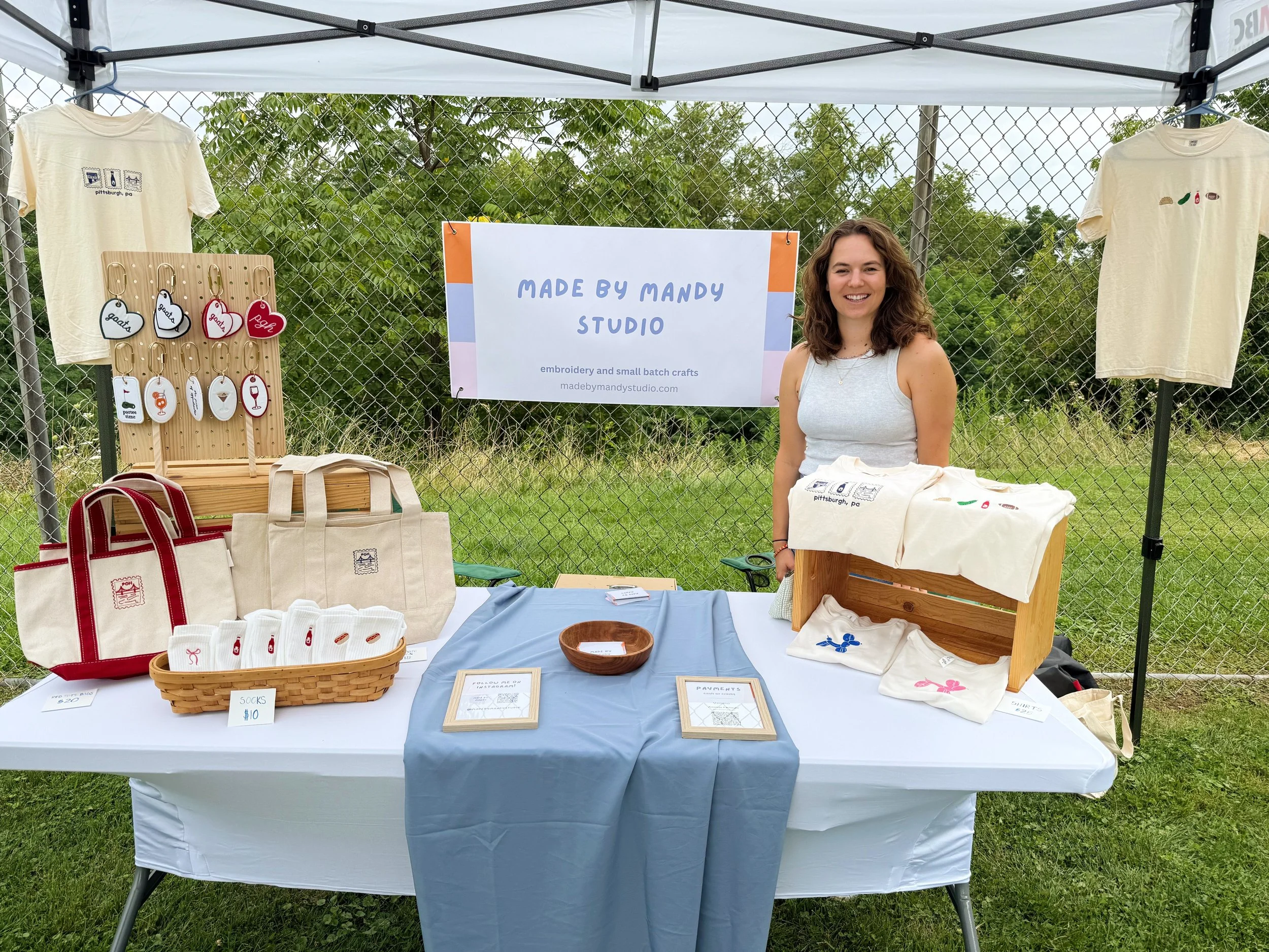 Outdoor booth displaying personalized embroidered items including T-shirts, tote bags, and keychains. A woman is standing behind the table, smiling. There is a white sign with purple text that reads 'Made by Mandy Studio' and indicates they do embroidery and small batch crafts.