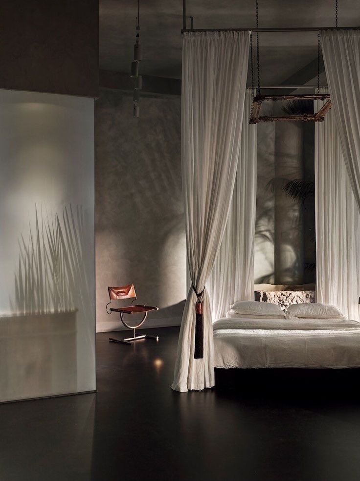 A bedroom with white curtains around the bed, a white bed with pillows, and a modern brown leather chair with a chrome base in the corner. The scene is softly lit, creating shadows and a calm ambiance.