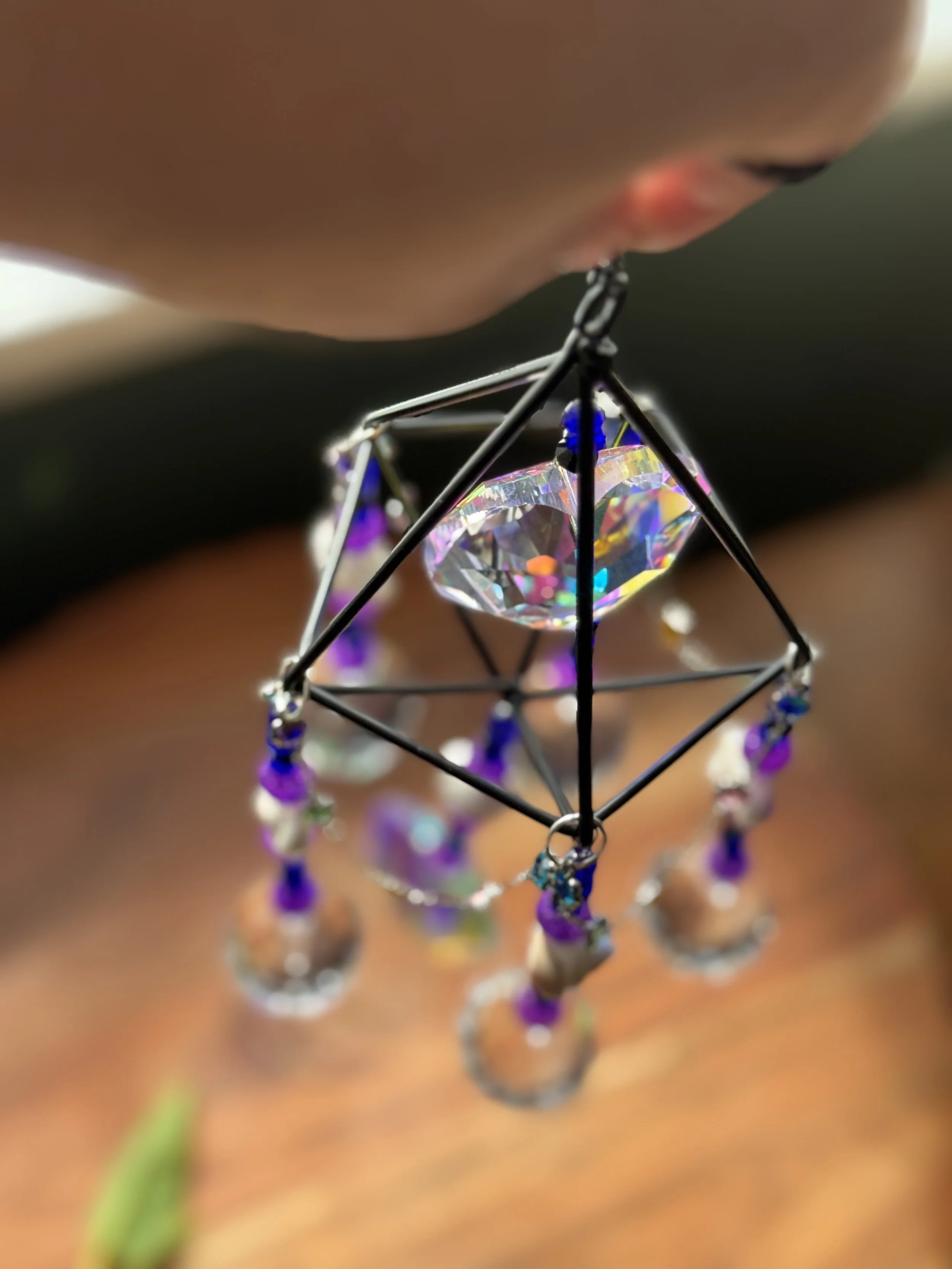 Close-up of a person holding a geometric metal and crystal ornament with purple beads and rings, hanging from their fingers.