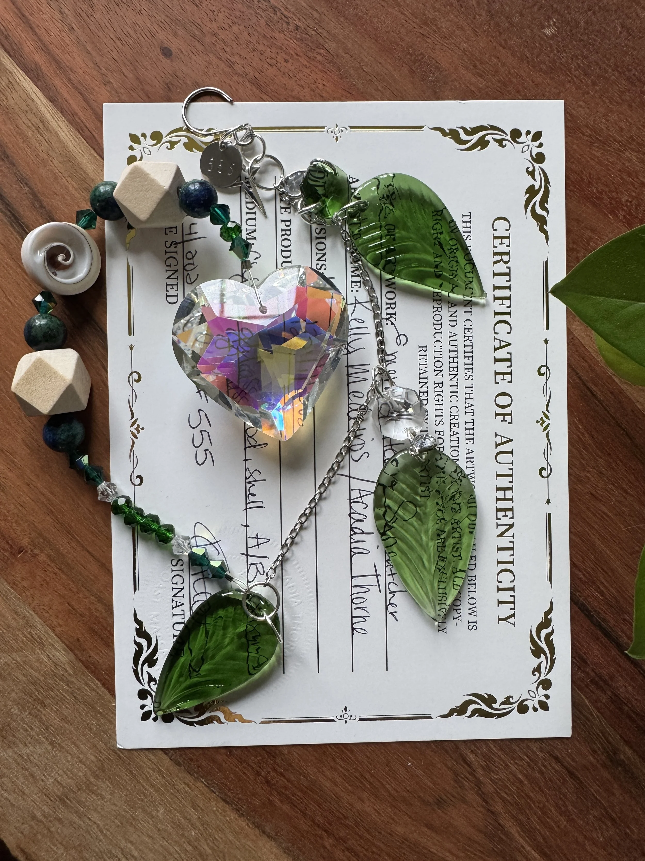 A certificate of authenticity with several pieces of jewelry placed on top, including a large iridescent heart-shaped pendant, three green leaf-shaped glass pendants, a shell-shaped charm, and a beaded bracelet with various colored beads.