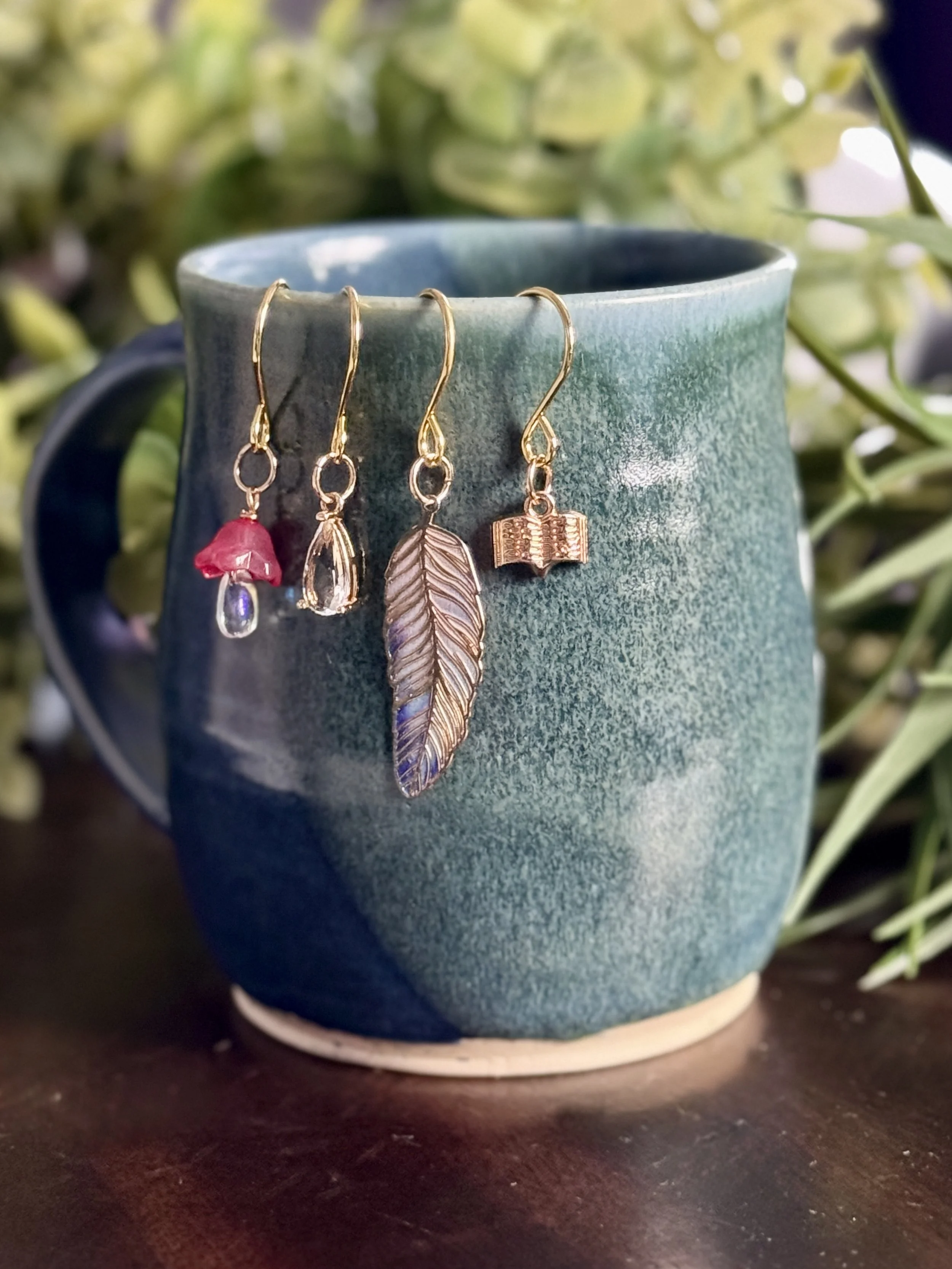 6th Alternative front view of a pink mushroom + book + feather + crystal plant charms hanging on a mug with leafy background