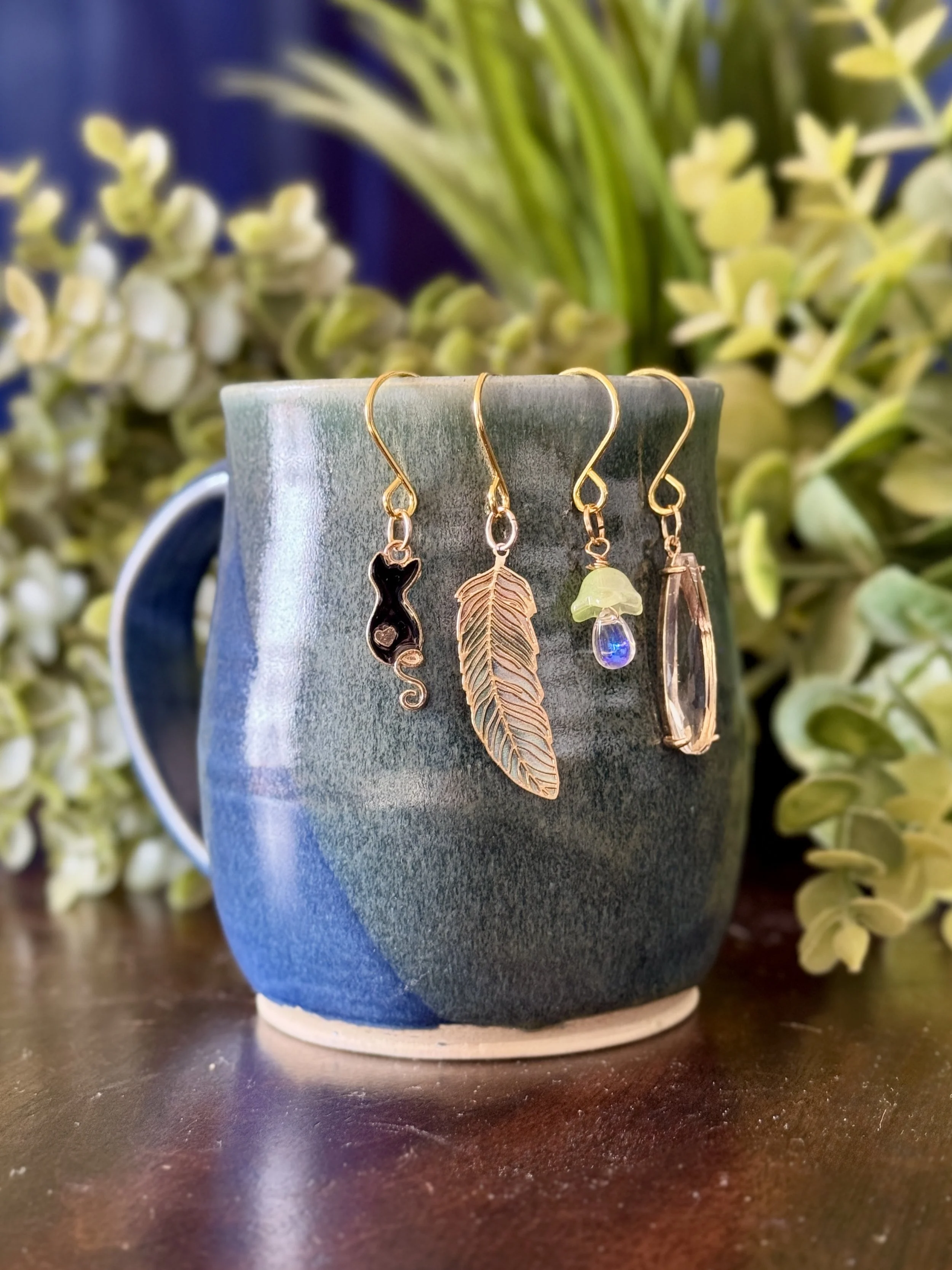 5th Front View of Black cat + green mushroom + green feather + crystal pendant plant charm set on blue mug with leafy background