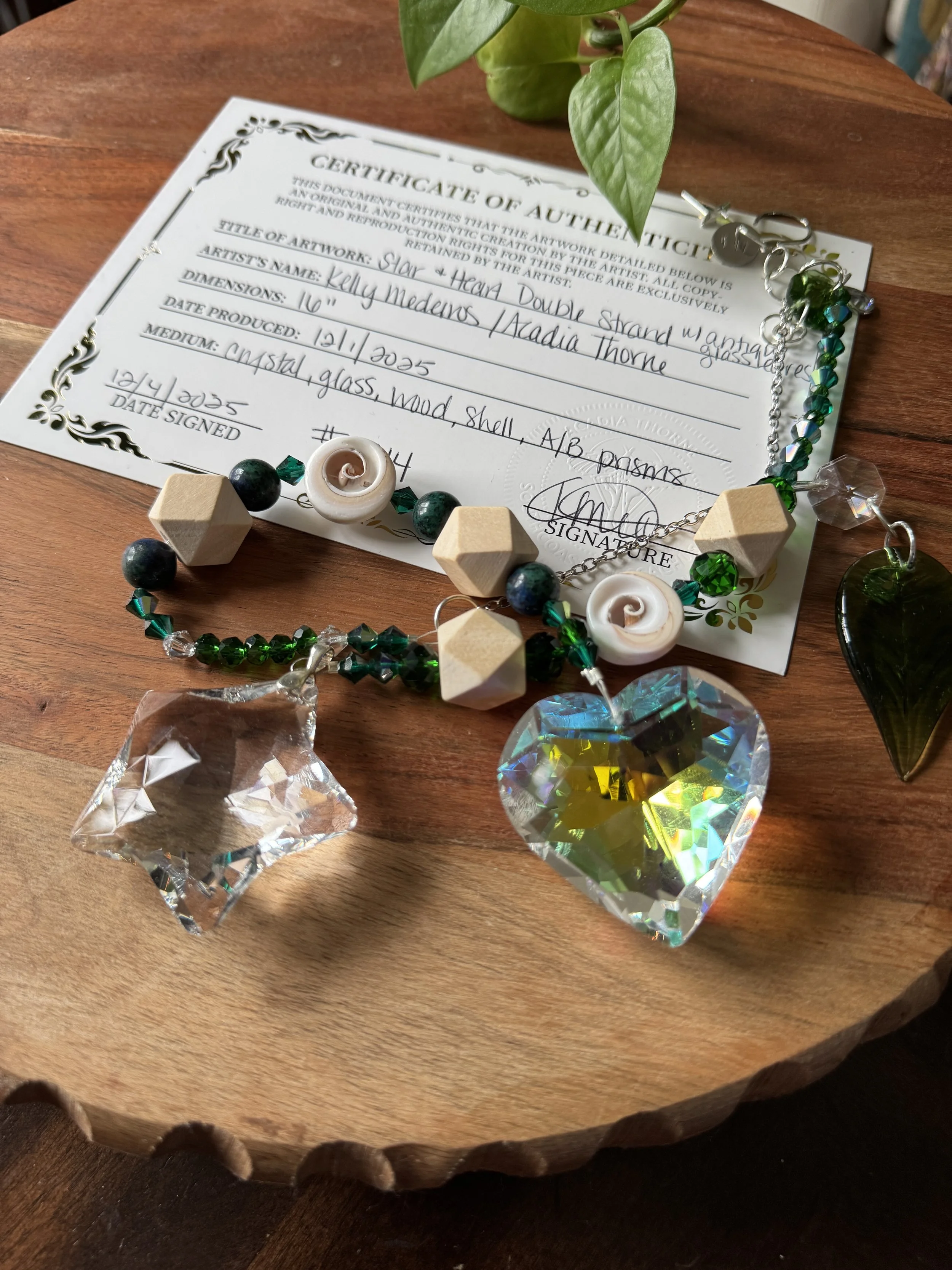 Colorful handcrafted beaded necklace with a large iridescent heart pendant, other small crystal pendants, and a certificate of authenticity on a wooden surface with a green plant nearby.