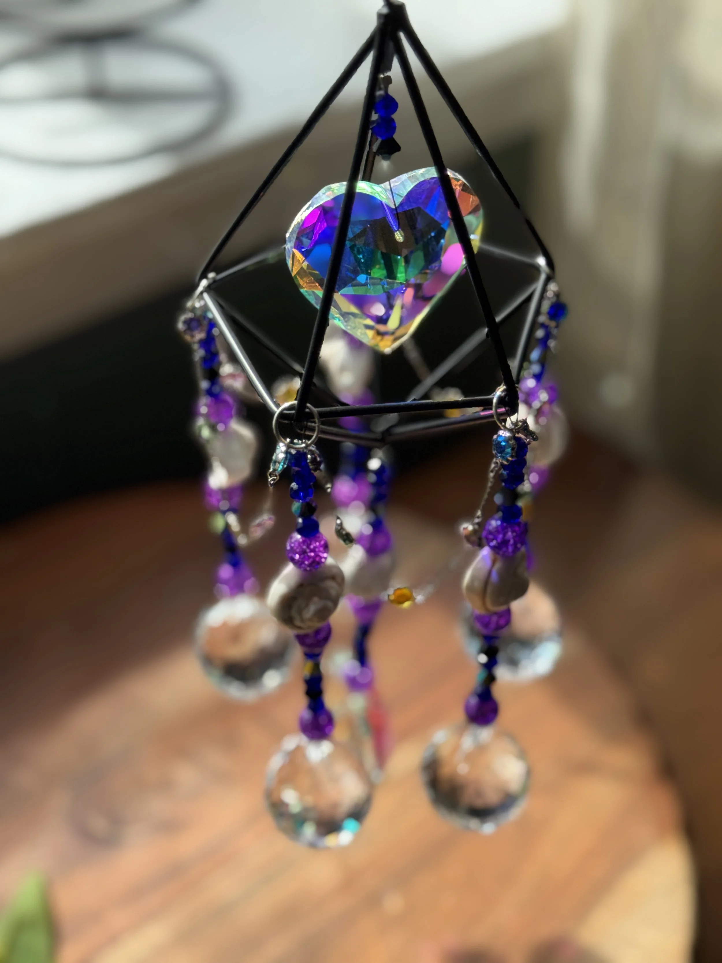 A colorful, iridescent heart-shaped crystal hanging inside a geometric metal frame, decorated with purple, blue, and clear beads.