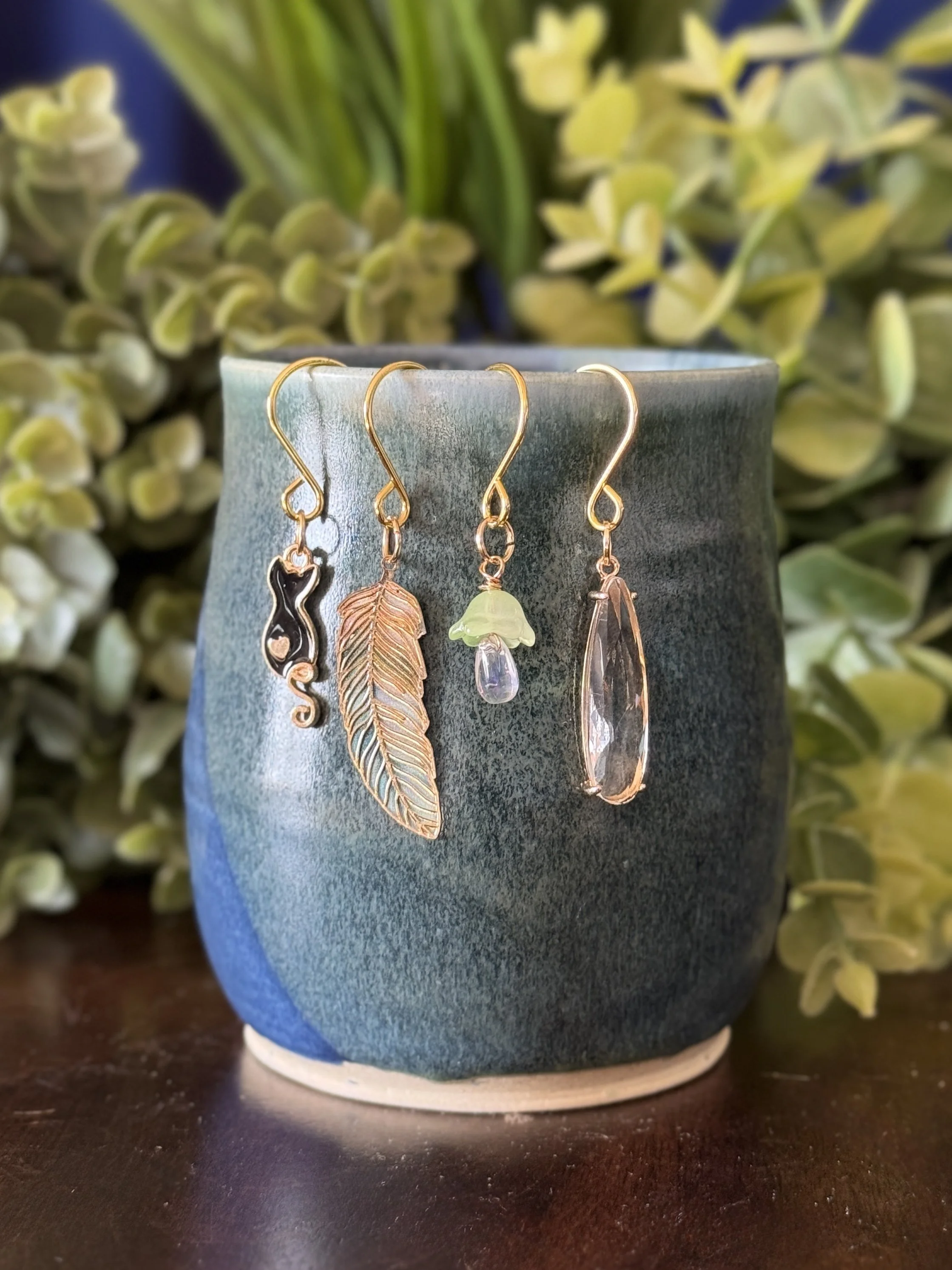 2nd Front View of Black cat + green mushroom + green feather + crystal pendant plant charm set on blue mug with leafy background