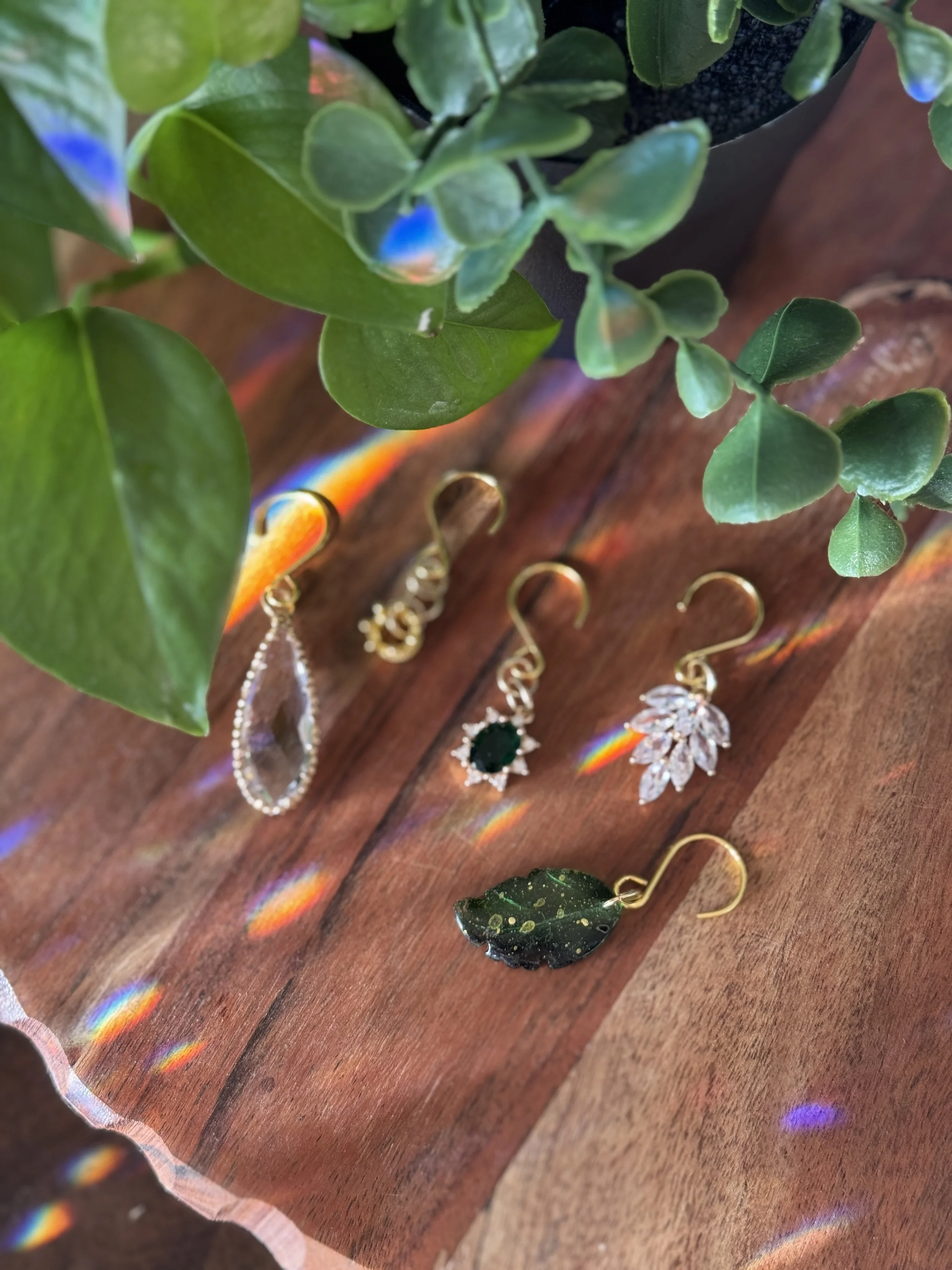 Four pairs of earrings with different designs laid on a wooden surface, with a potted plant with green leaves in the background, and colorful light refraction creating rainbow effects.