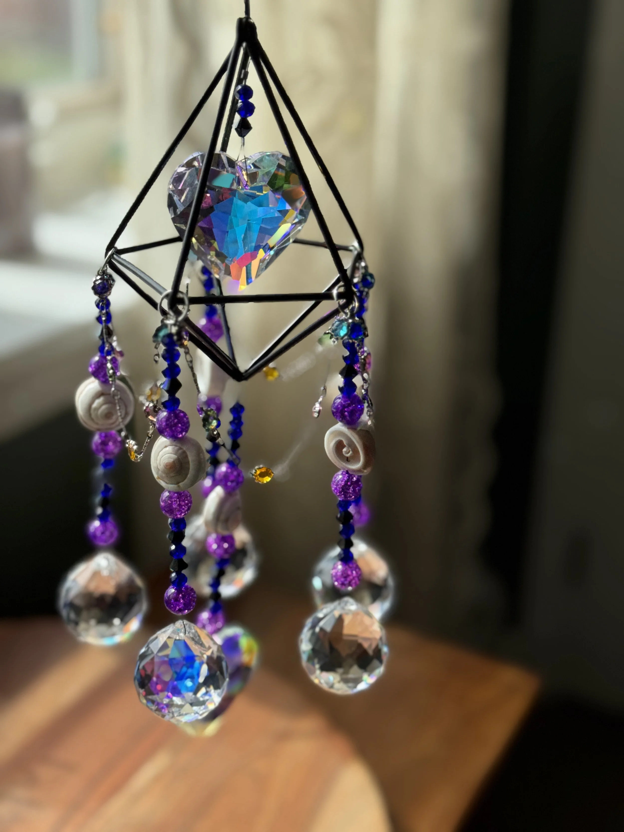 A decorative hanging ornament featuring a central iridescent gemstone heart, surrounded by dangling purple, clear, and iridescent beads and shells, suspended from a wire frame.