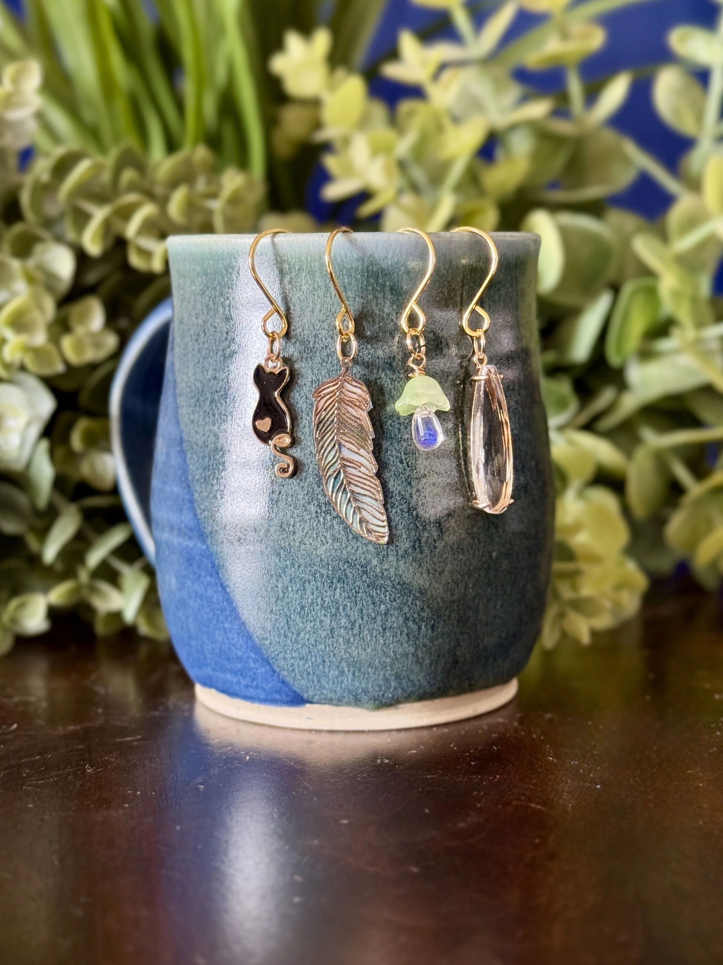 7th Front View of Black cat + green mushroom + green feather + crystal pendant plant charm set on blue mug with leafy background