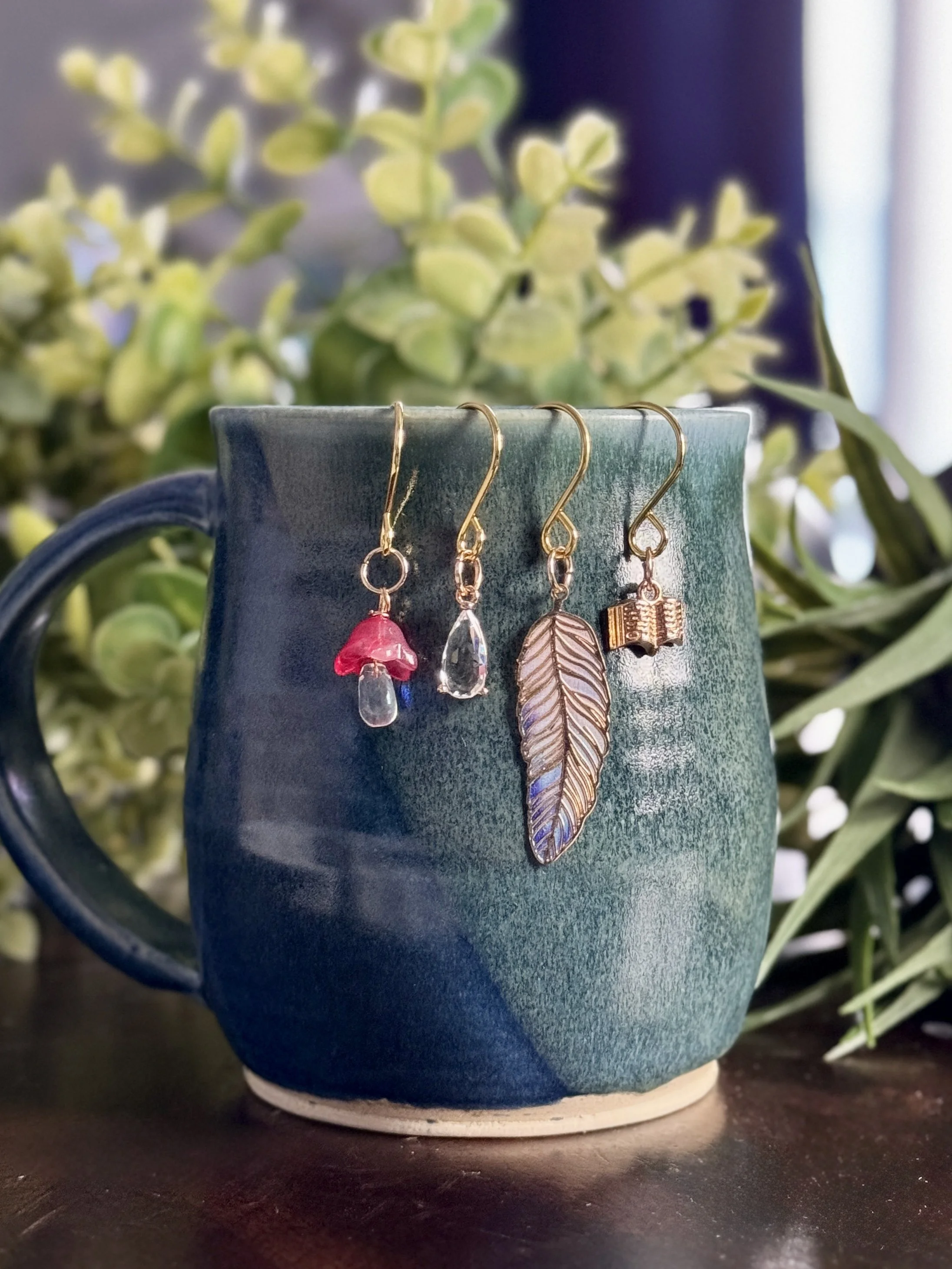 3rd Alternative front view of a pink mushroom + book + feather + crystal plant charms hanging on a mug with leafy background