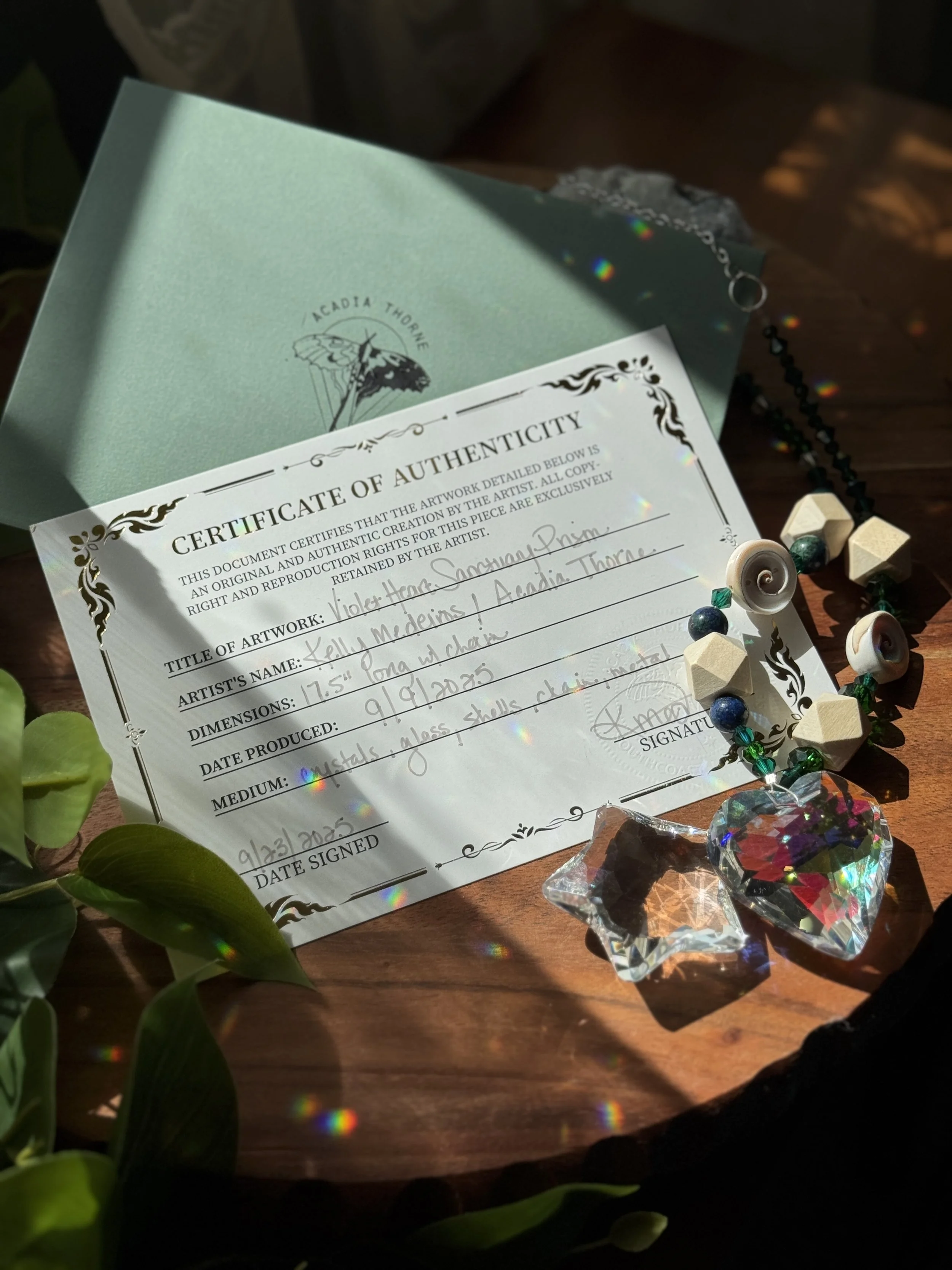A certificate of authenticity for a piece of artwork, a beaded necklace with shell and glass beads, a glass heart, and a smaller glass star-shaped ornament on a wooden surface, with sunlight creating rainbow reflections.