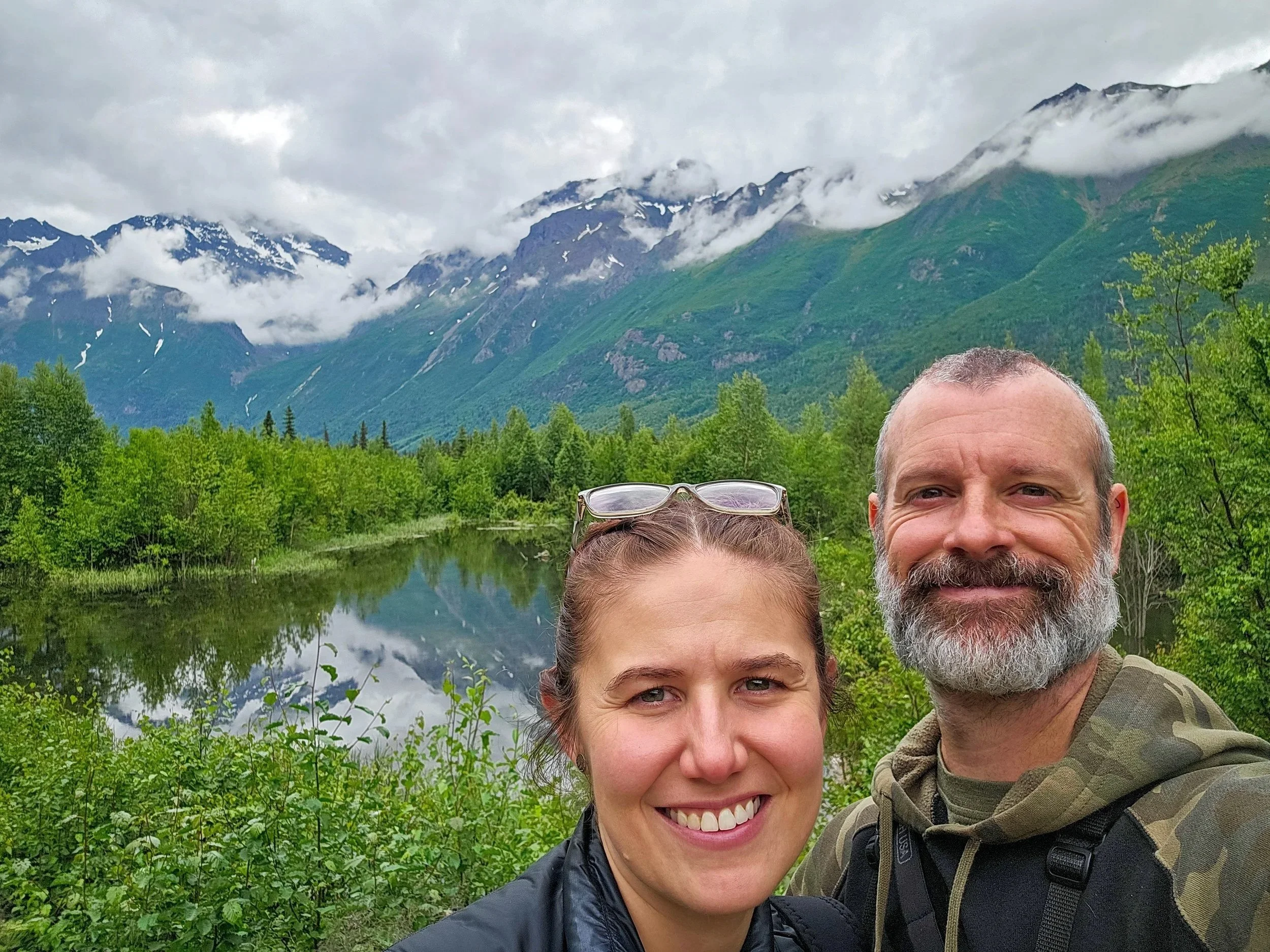 Michelle and Chris Coverdill taking a selfie at Eagle River Nature Center with lush green trees, a calm river, and mountain peaks with patches of snow in the background.