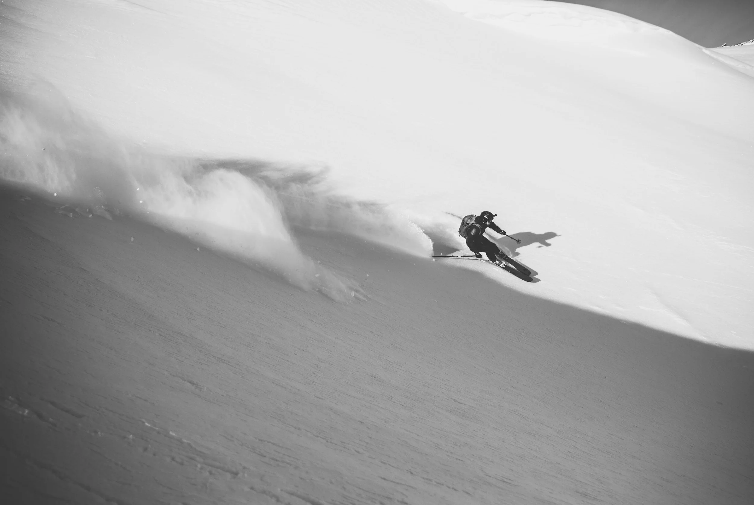 A skier in black gear descending a snow-covered mountain slope, creating a spray of snow behind them.