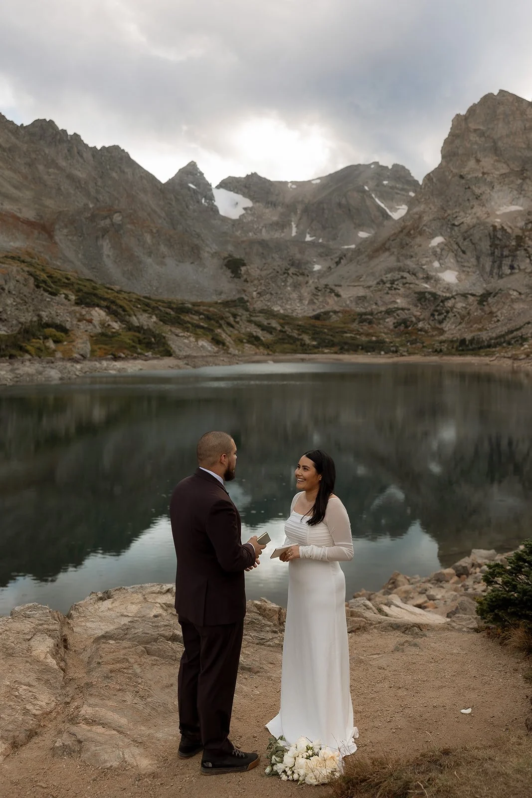 A couple is exchanging vows in front of a serene mountain lake, surrounded by rocky terrain and rugged peaks. The bride is wearing a white dress and holding flowers, while the groom is dressed in a dark suit, both appearing happy and engaged in the m