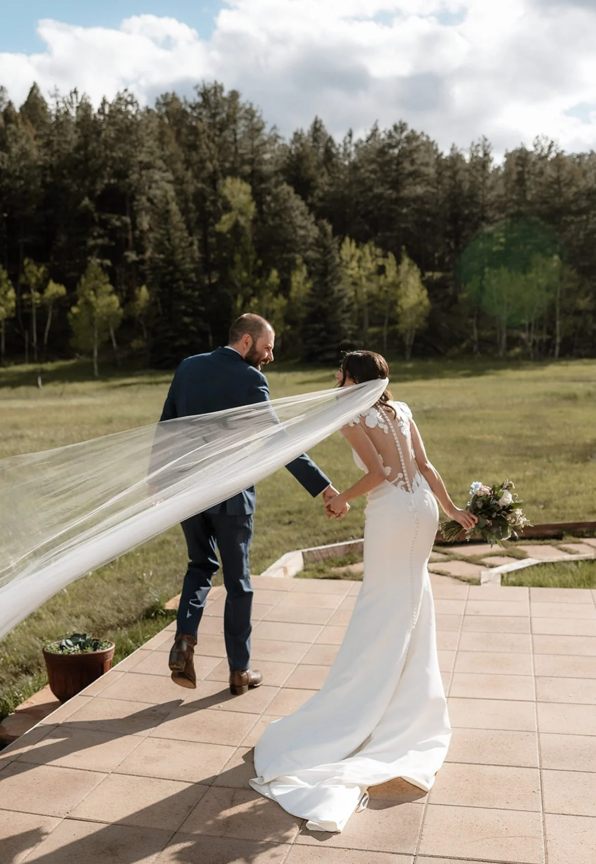 Bride and groom holding hands, walking outdoors with bride's veil flowing, surrounded by lush greenery and forest.